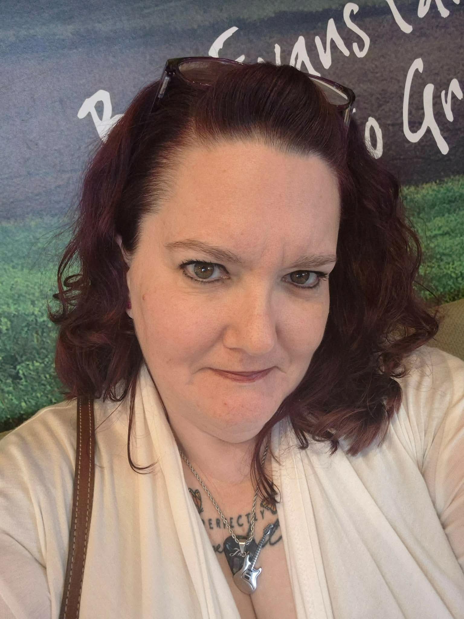 Woman with curly red hair wearing glasses on her head, a necklace with a guitar pendant, and a beige top, standing in front of a background with greenery and white text.