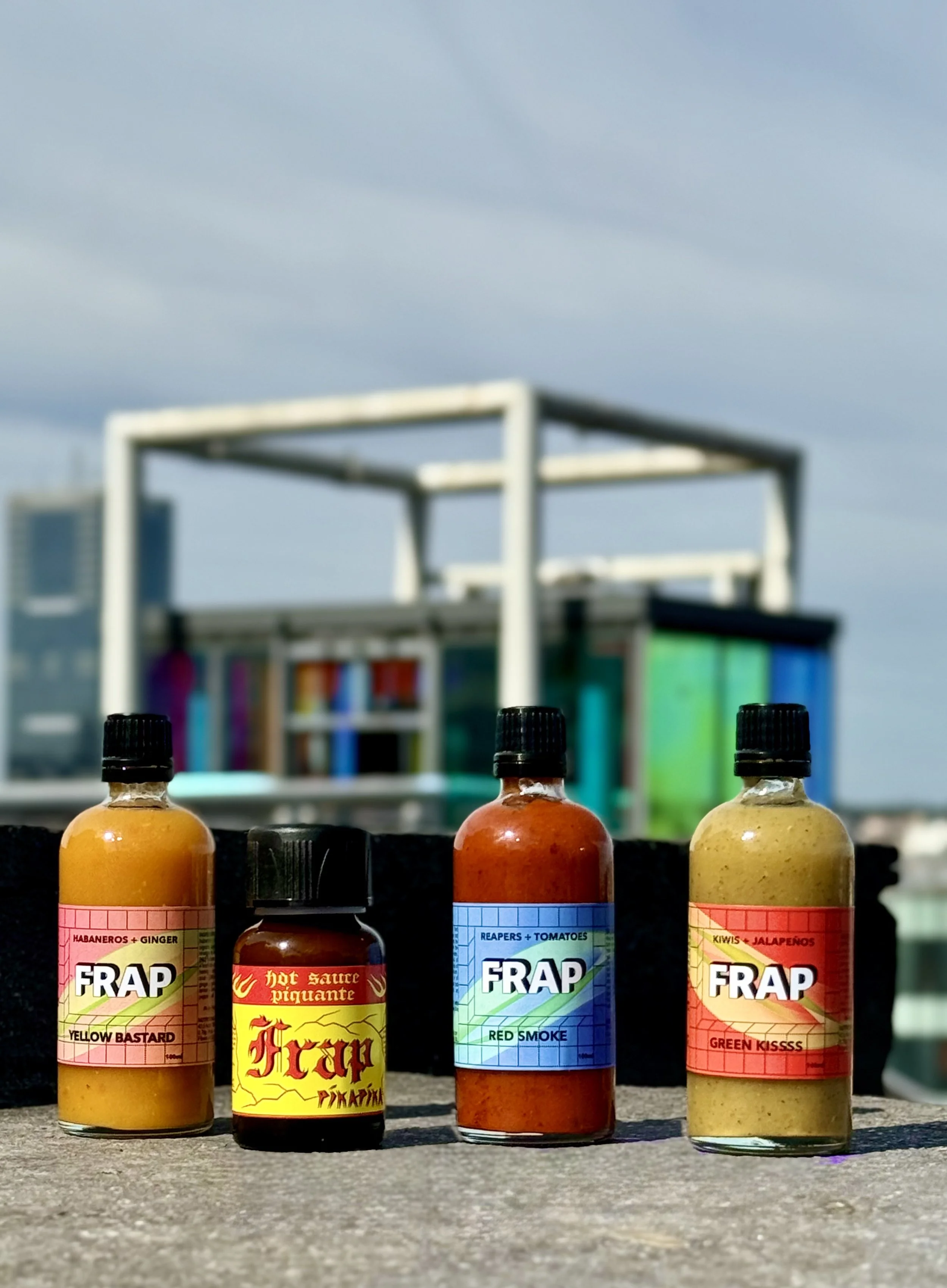 Quatre bouteilles de sauces piquantes avec des étiquettes colorées, placées sur une surface en extérieur avec un bâtiment en arrière-plan.