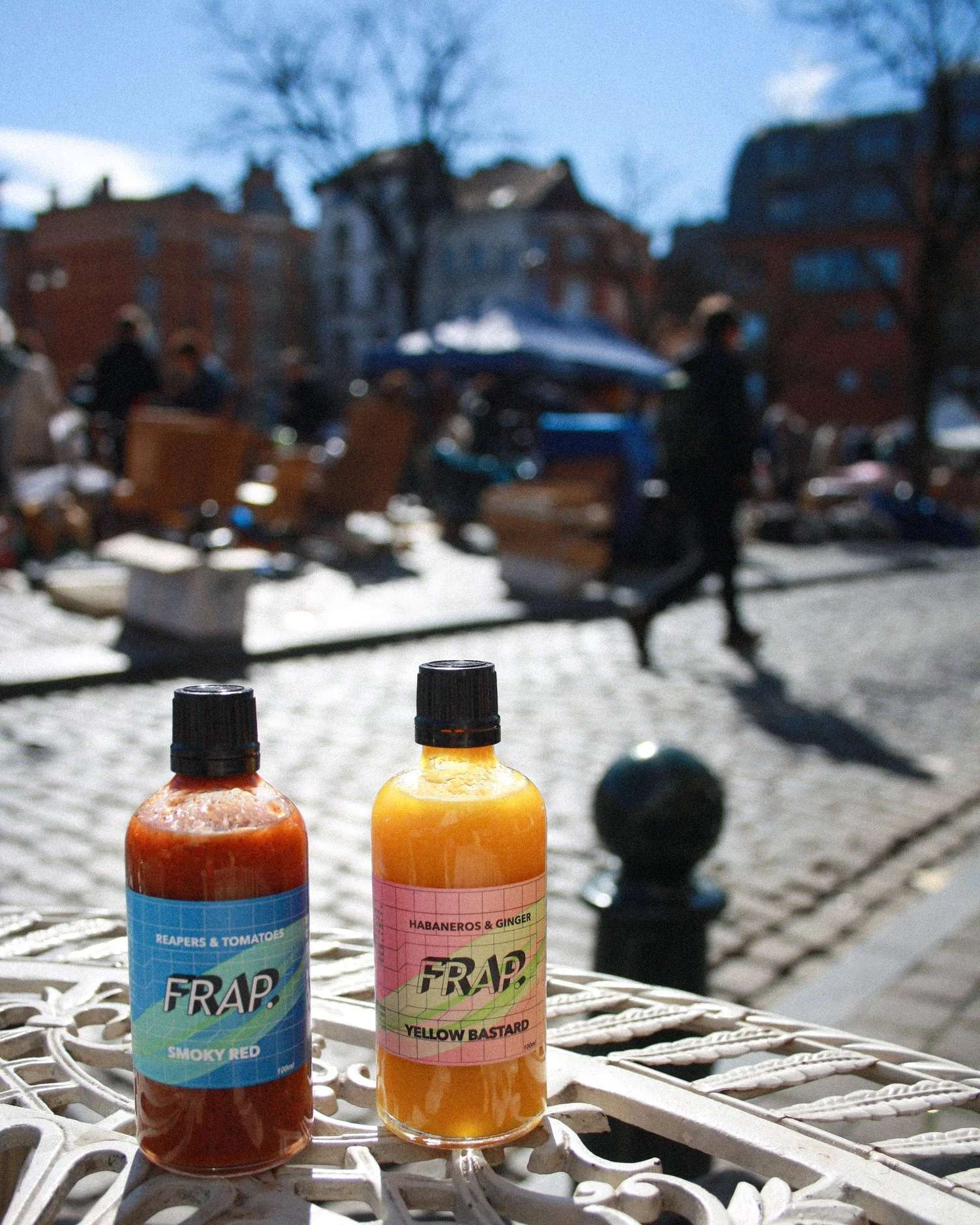 Deux bouteilles de liquide de couleur orange et rouge avec des étiquettes, placées sur une surface en métal, avec un marché en plein air en arrière-plan et des personnes qui marchent.