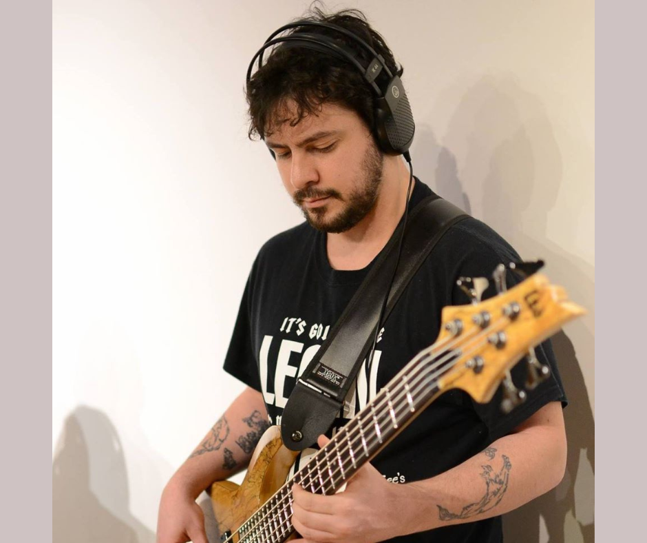 A voice actor for the horror fiction podcast The White Vault. A man with a black tee-shirt and a wooden 5-string bass guitar. He wears headphones and has curly brown hair and a short beard. He also has tattoos on both arms.