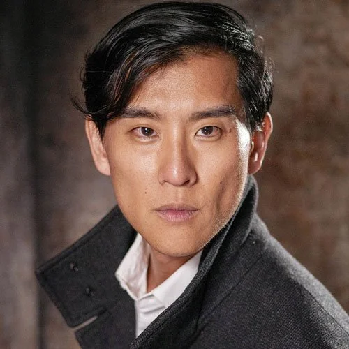 A voice actor from the VAST Horizon science fiction podcast. A man with combed black hair and a formal grey overcoat that sits over a white collared shirt. He looks intense but friendly.