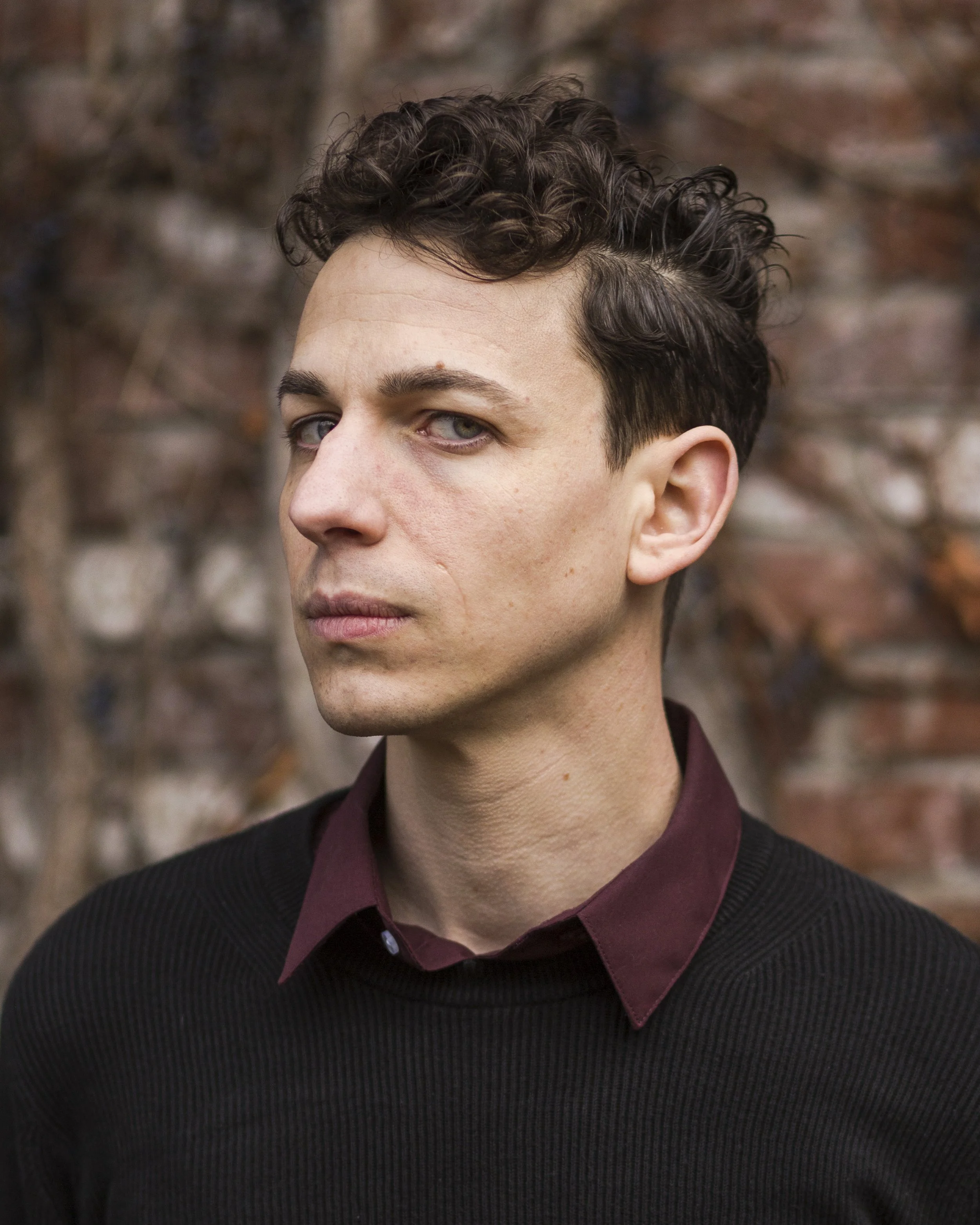A man with curly brown hair and a purple formal shirt under a black sweater. He's scowling. This is Travis Vengroff, the sound designer and producer of horror fiction podcasts.