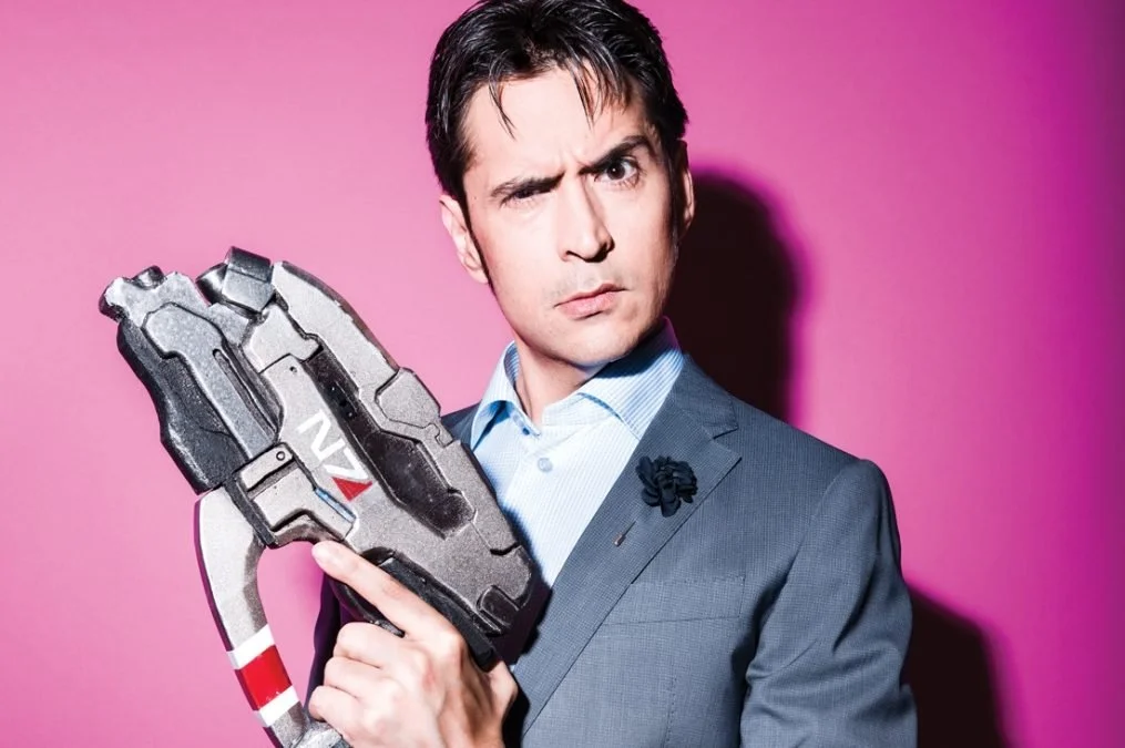 An actor for Don't Mind Sealskin Rock, a horror mystery fiction podcast. A man with a dark clean haircut and a suit with a blue shirt, and a Stiletto Pistol from Mass effect that reads N7. He makes a silly face by raising one eyebrow.