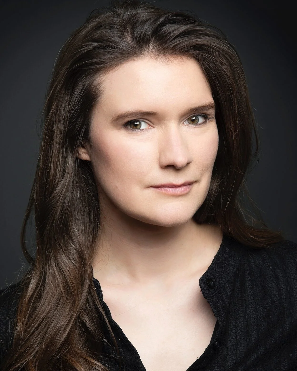 A voice actor for the horror fiction podcast The White Vault. A woman with long brown hair and brown eyes. Her expression is mysterious and she wears a black formal top..