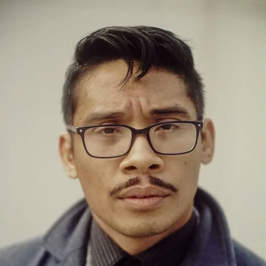 An actor for Don't Mind Cruxmont, a mystery fiction podcast. A man with a thin mustache, black rectangular glasses, and a very formal coat and button up shirt with a tie. His hair is very short on the sides and styled on top. He looks professional.