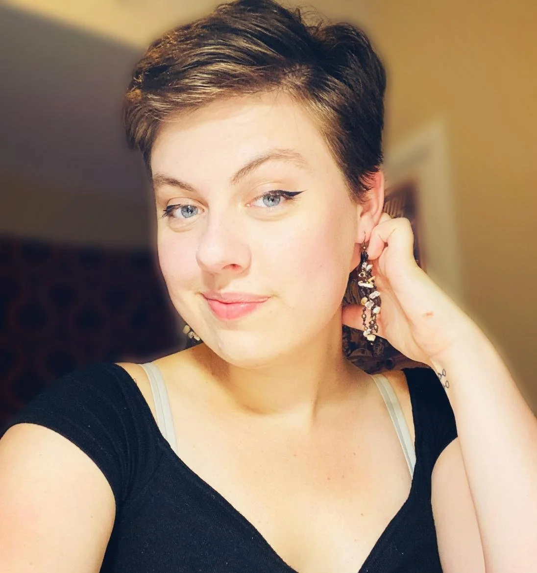 A voice actor for the horror fiction podcast The White Vault. A feminine person with short dark-blond hair and grey eyes smiles. They wear a black dress with stony earrings,  and one hand  is behind their head.