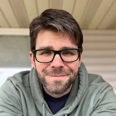 An actor for Don't Mind Sealskin Rock, a horror mystery fiction podcast. The most Canadian photo you can imagine: a bearded man on his porch with black glasses, a hoodie, and a friendly smile. His hair a grey and black.