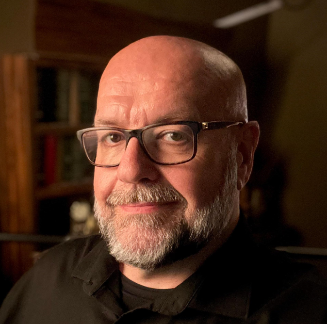 An actor from Liberty: Tales from the Tower, a horror fiction podcast. A bald man with a white beard and glasses. He wears a black formal shirt over a black undershirt. He is the podfather.