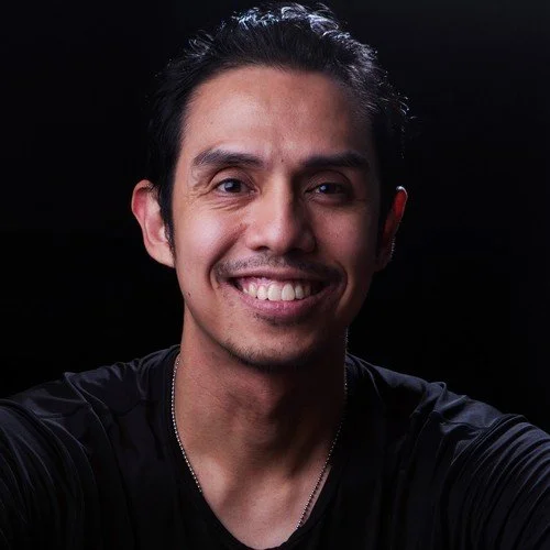 A voice actor for the horror fiction podcast The White Vault. A smiling man with a stubble mustache and black hair that is combed back. He smiles and wears a black shirt that looks super comfortable.