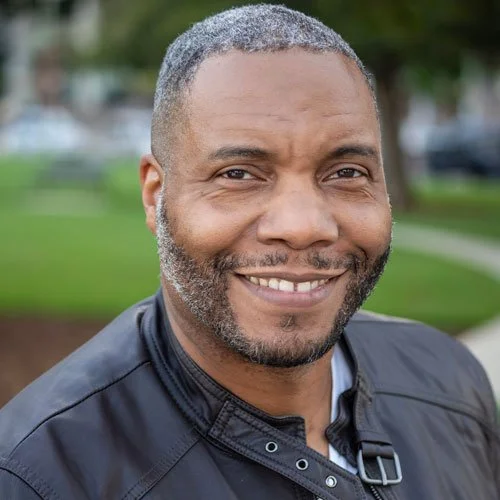 A voice actor from the VAST Horizon science fiction podcast. A man with short black and grey peppered hair and a short beard. He wears a black leather jacket that looks formal, and stands in front of a park.