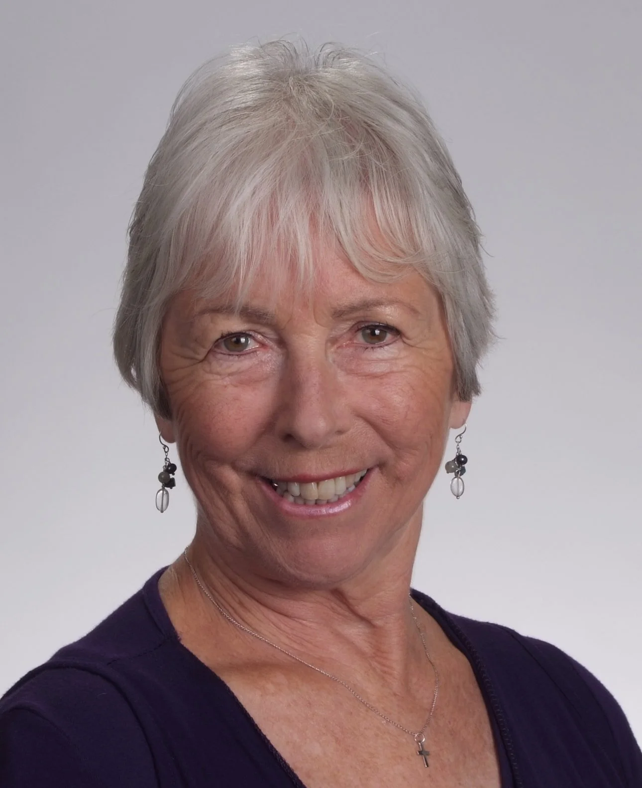 An actor for Don't Mind Cruxmont, a mystery fiction podcast. A smiling woman with ear-length white hair and cute earrings of circular stone and wire design. She has hazel eyes.
