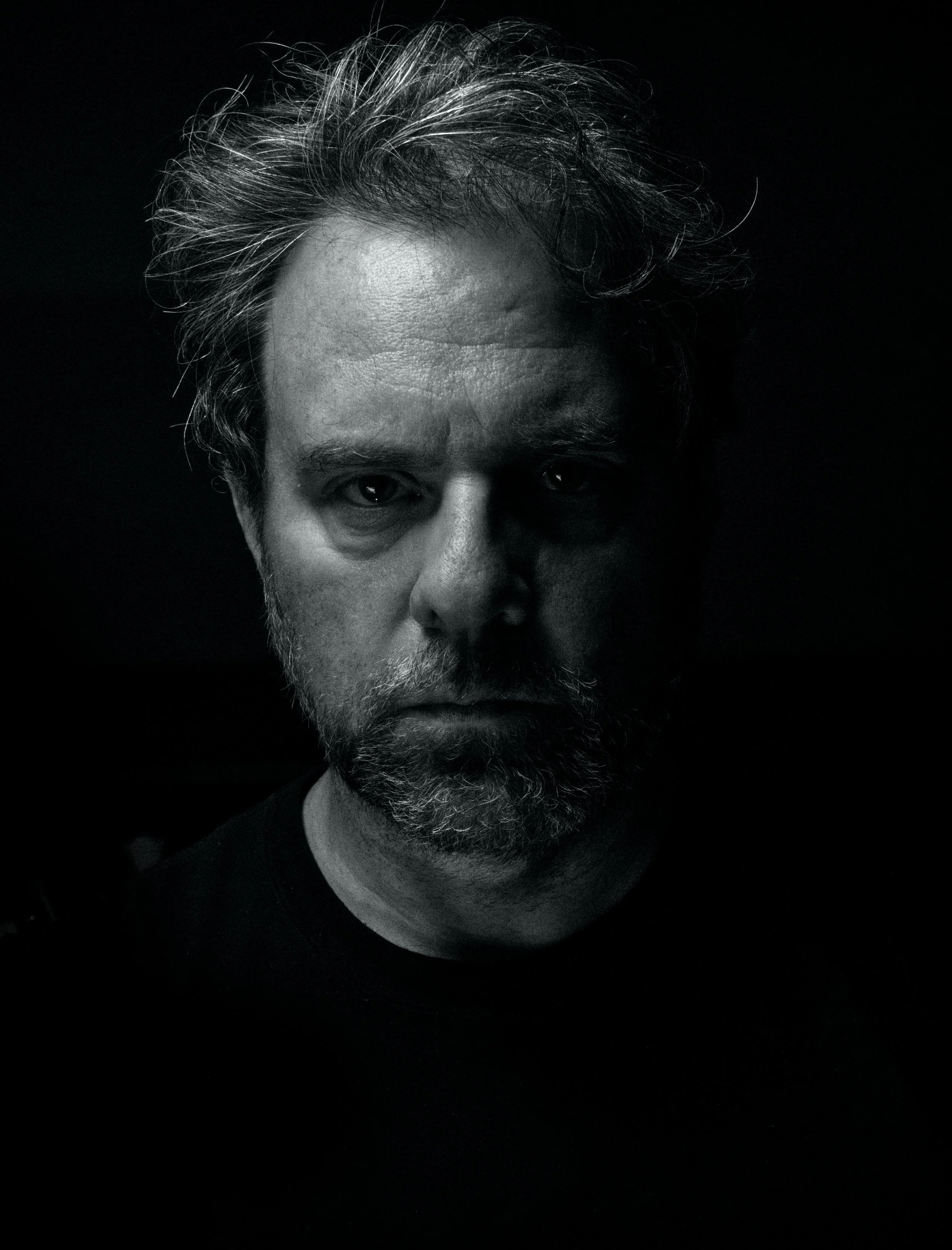 A voice actor for the horror fiction podcast The White Vault. A black and white photo of a man with a black shirt under dynamic lighting who has the look of wild genius. His unkempt hair and short beard lend to his intense expression.