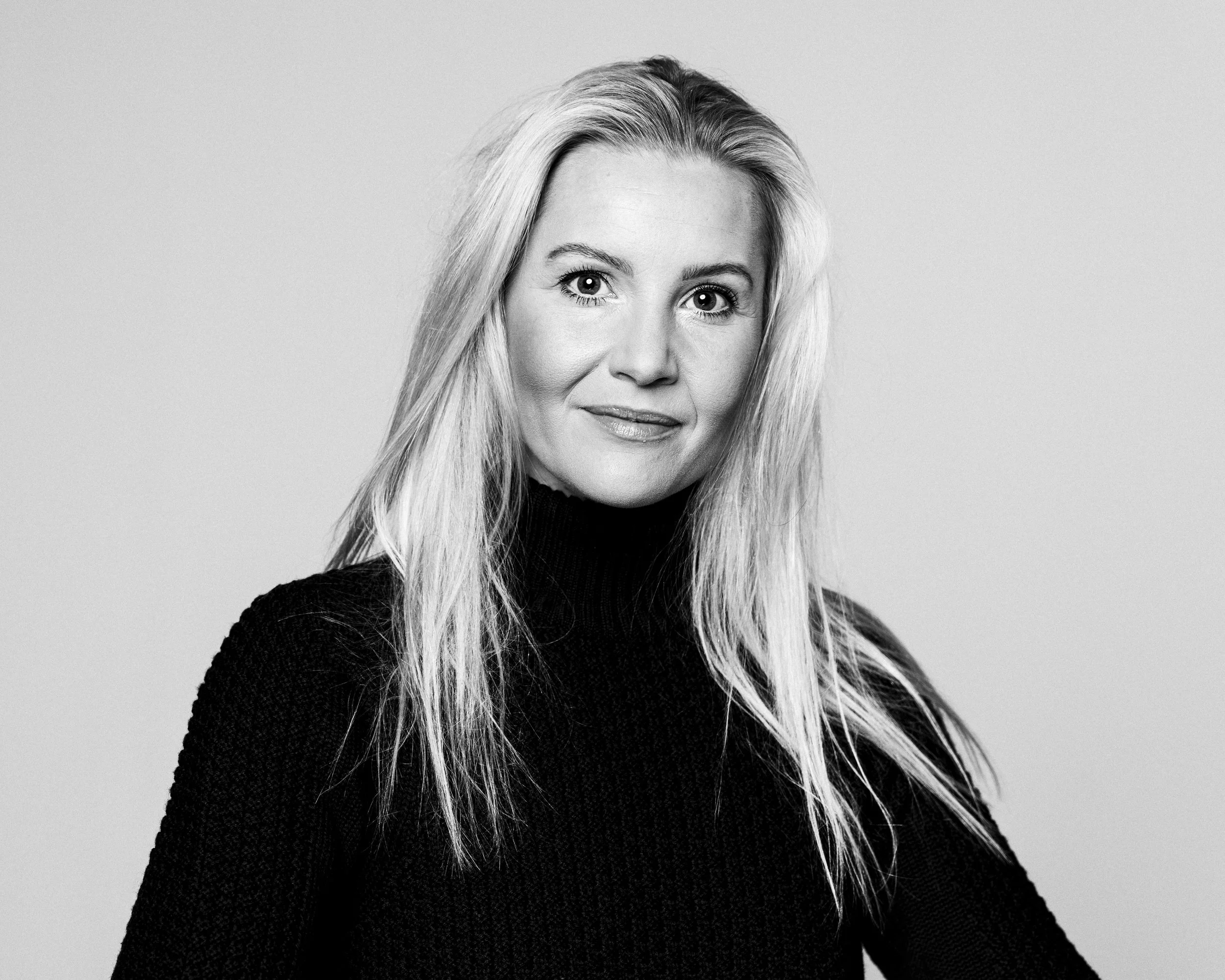 A voice actor for the horror fiction podcast The White Vault. A black and white image of a blond woman in a black sweater. Her hair extends past her shoulders and she has the look of a true stage professional.