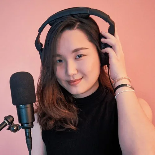 A voice actor from The Boar Knight, an all-ages fiction podcast. A woman in a black top, with headphones and a microphone. We shears 3 bracelets and has dark hair that extends past her shoulders. She smiles mysteriously.