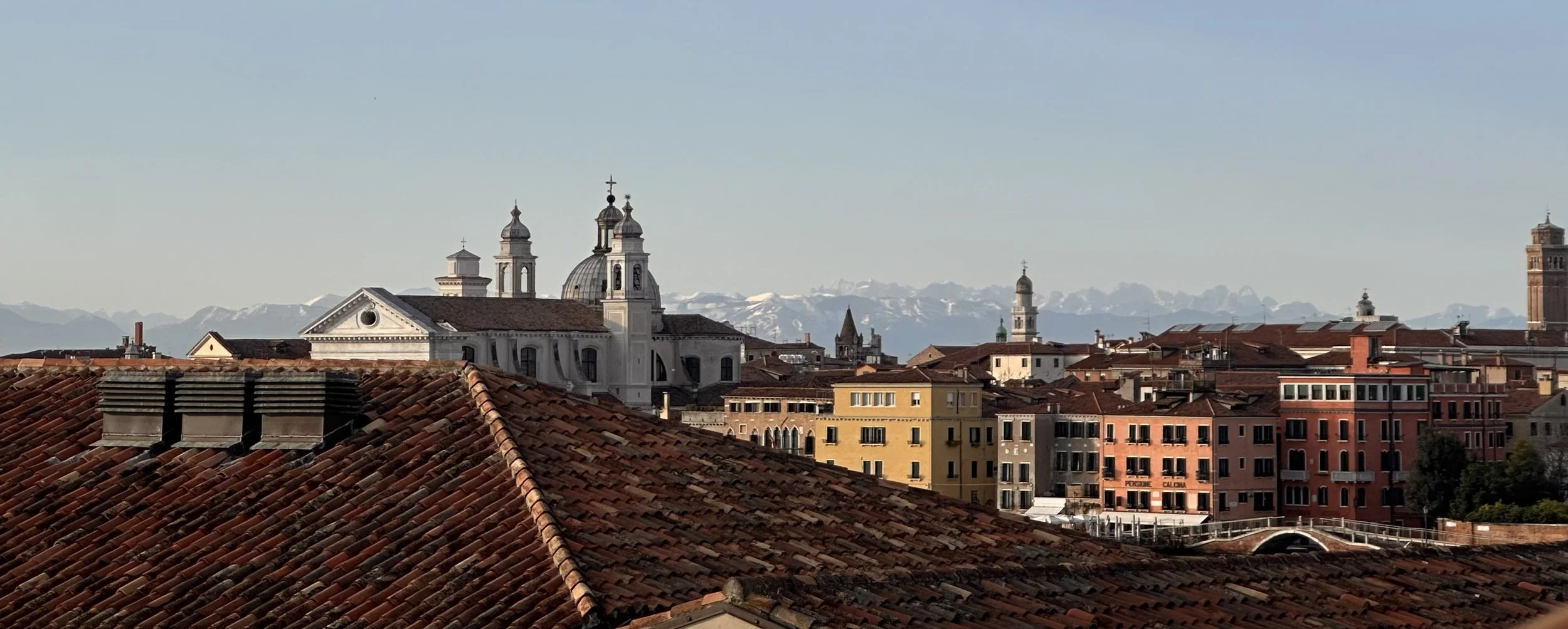 Panorama della città con tetti di tegole rosse e edifici storici, montagne innevate sullo sfondo.