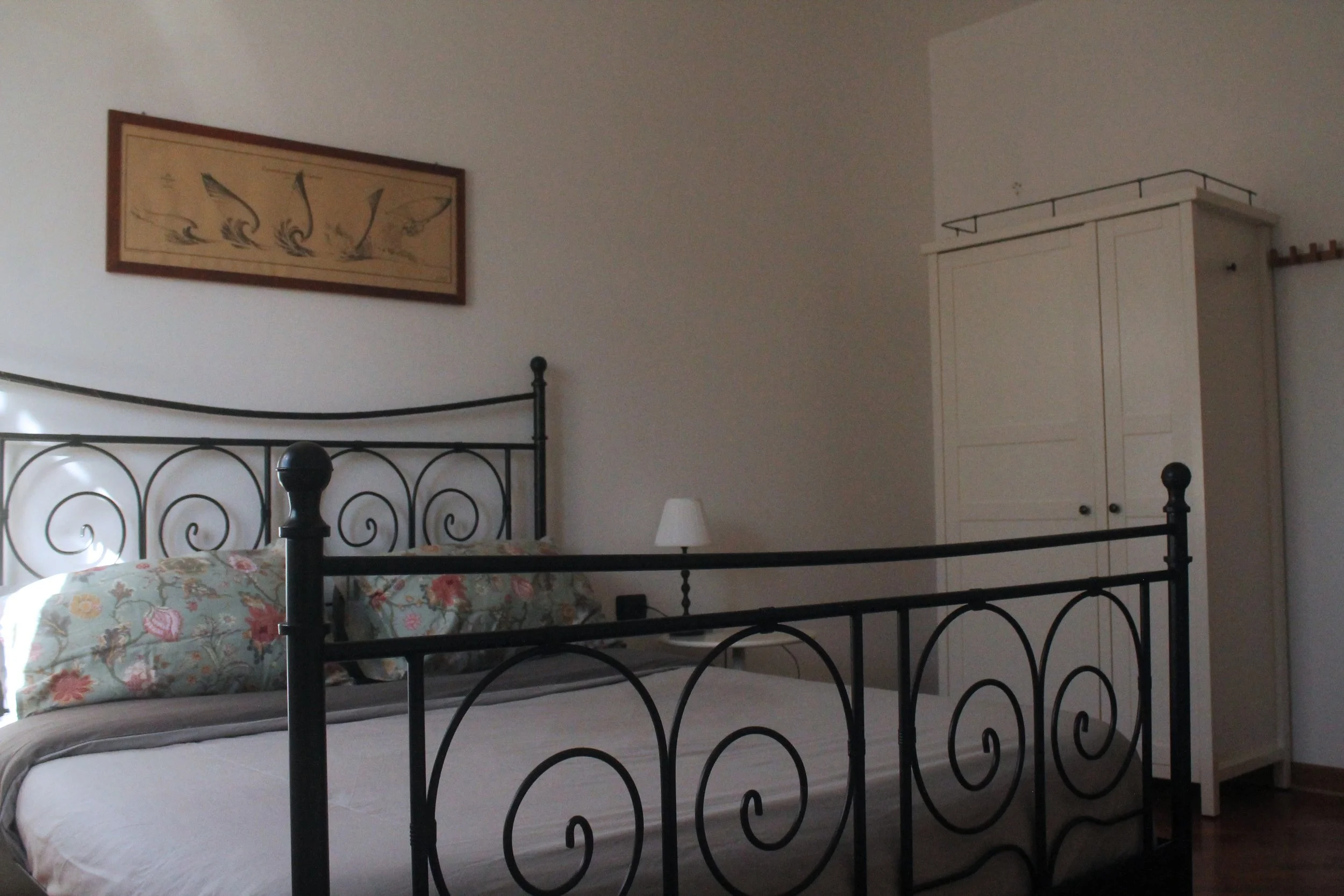 Camera da letto con letto in ferro battuto nero, biancheria da letto chiara, due cuscini con copertina floreale, parete con quadro con disegni stilizzati di uccelli, lampada da notte bianca e armadio bianco.