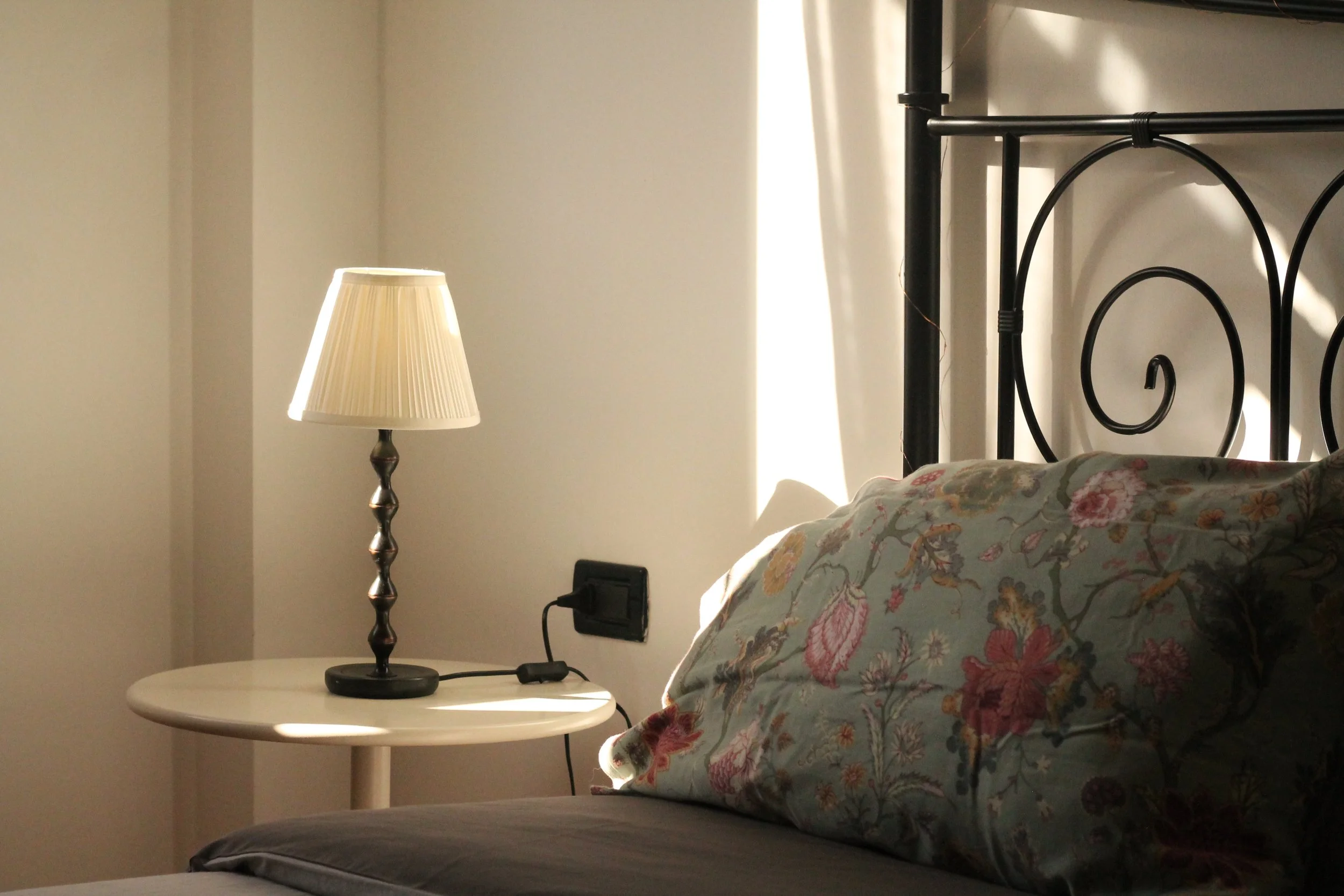 Letto con cuscino a fiori, lampada da tavolo accesa su comodino vicino alla testa del letto, luce naturale che entra dalla finestra.