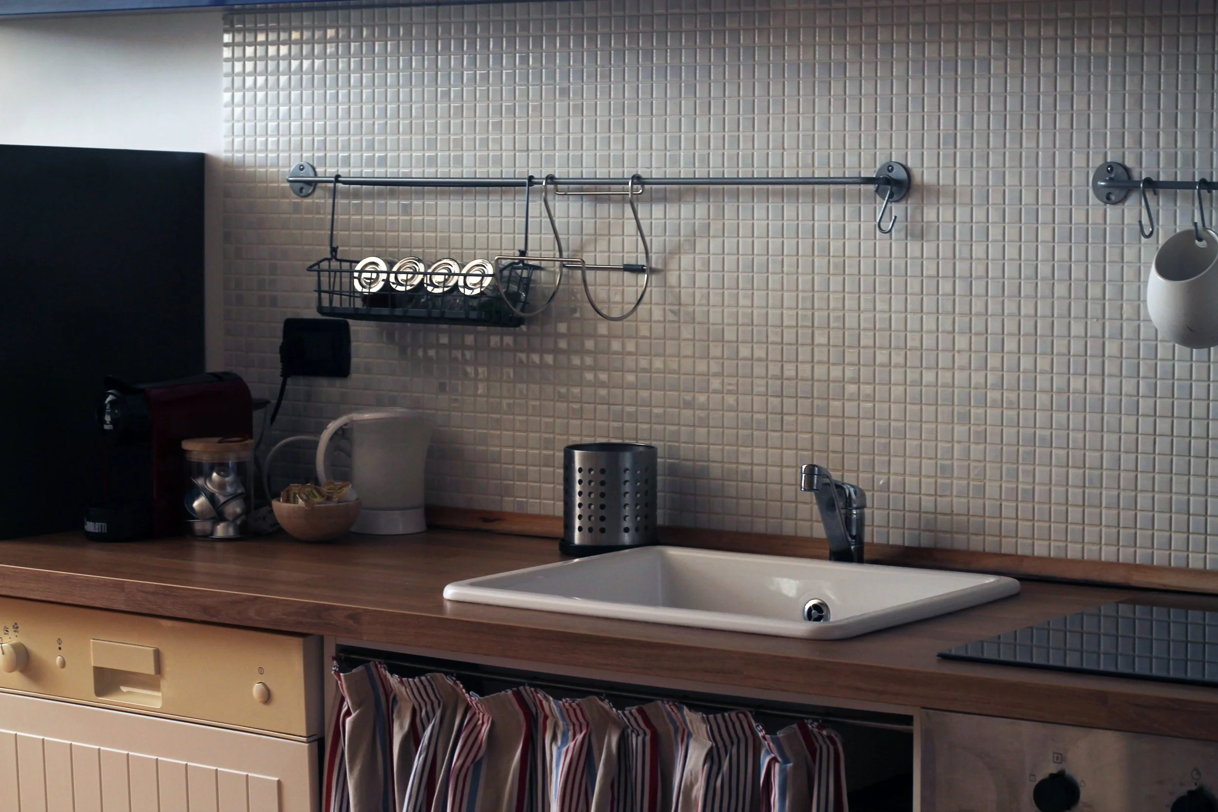 Angolo di cucina con lavandino, piano di lavoro in legno, stoviglie, macchina da caffè, tazzine, dispenser per utensili, mosaic tile bianco sulla parete, e vari elementi da cucina.