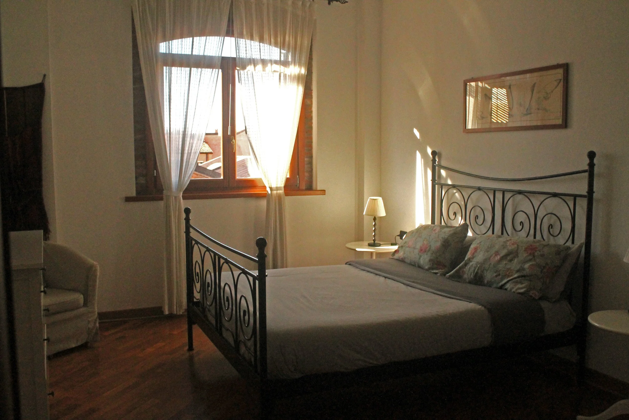 Letto matrimoniale con lenzuola e cuscini floreali, lampada da tavolo sul comodino, finestra con tende leggere, pareti chiare, quadro sopra il letto, seduta a sinistra, pavimento in legno.