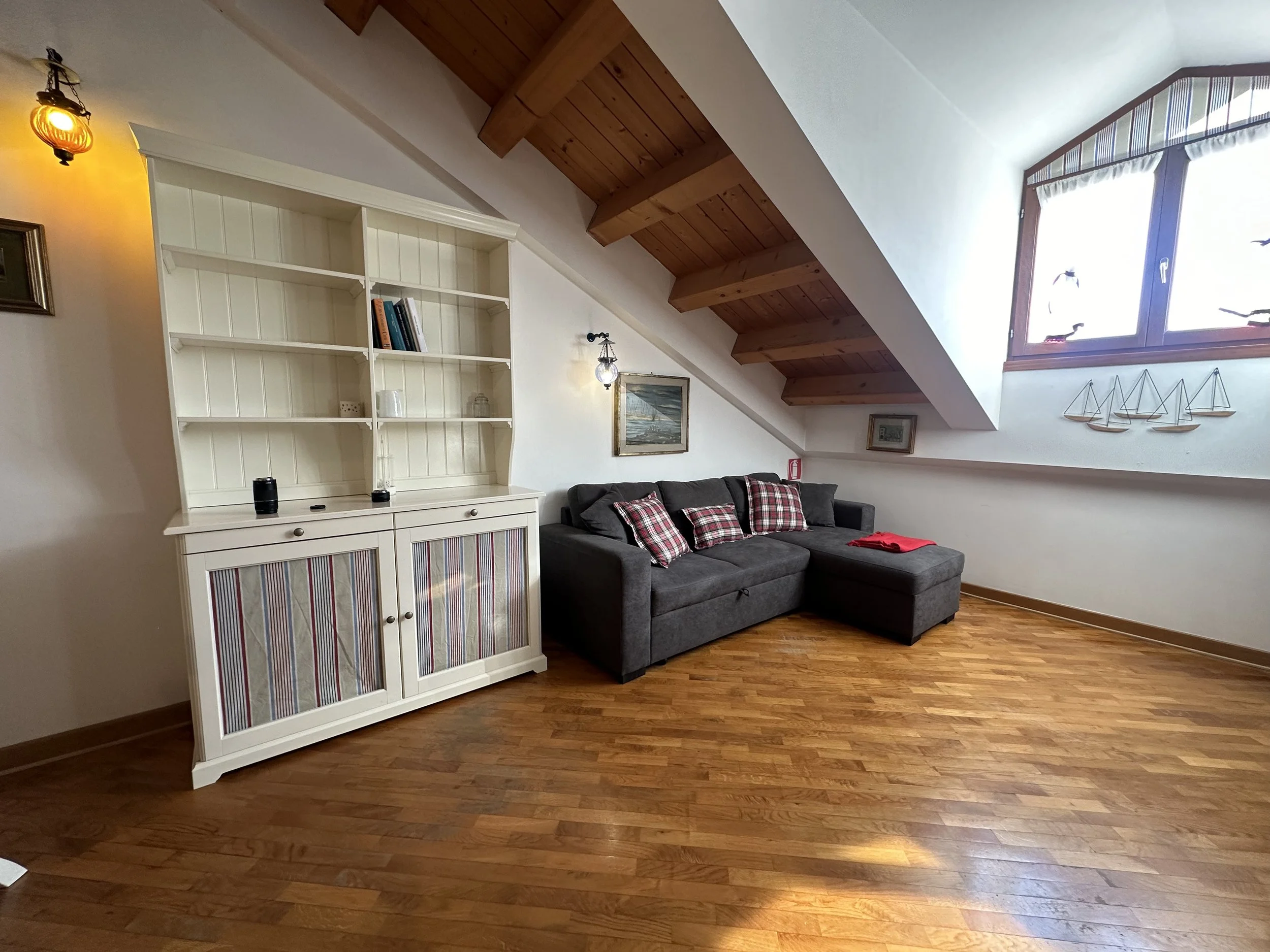 Soggiorno con divano grigio, tappetini a quadretti rosso e bianco, libreria bianca, pareti bianche, pavimento in legno e soffitto con travi di legno.