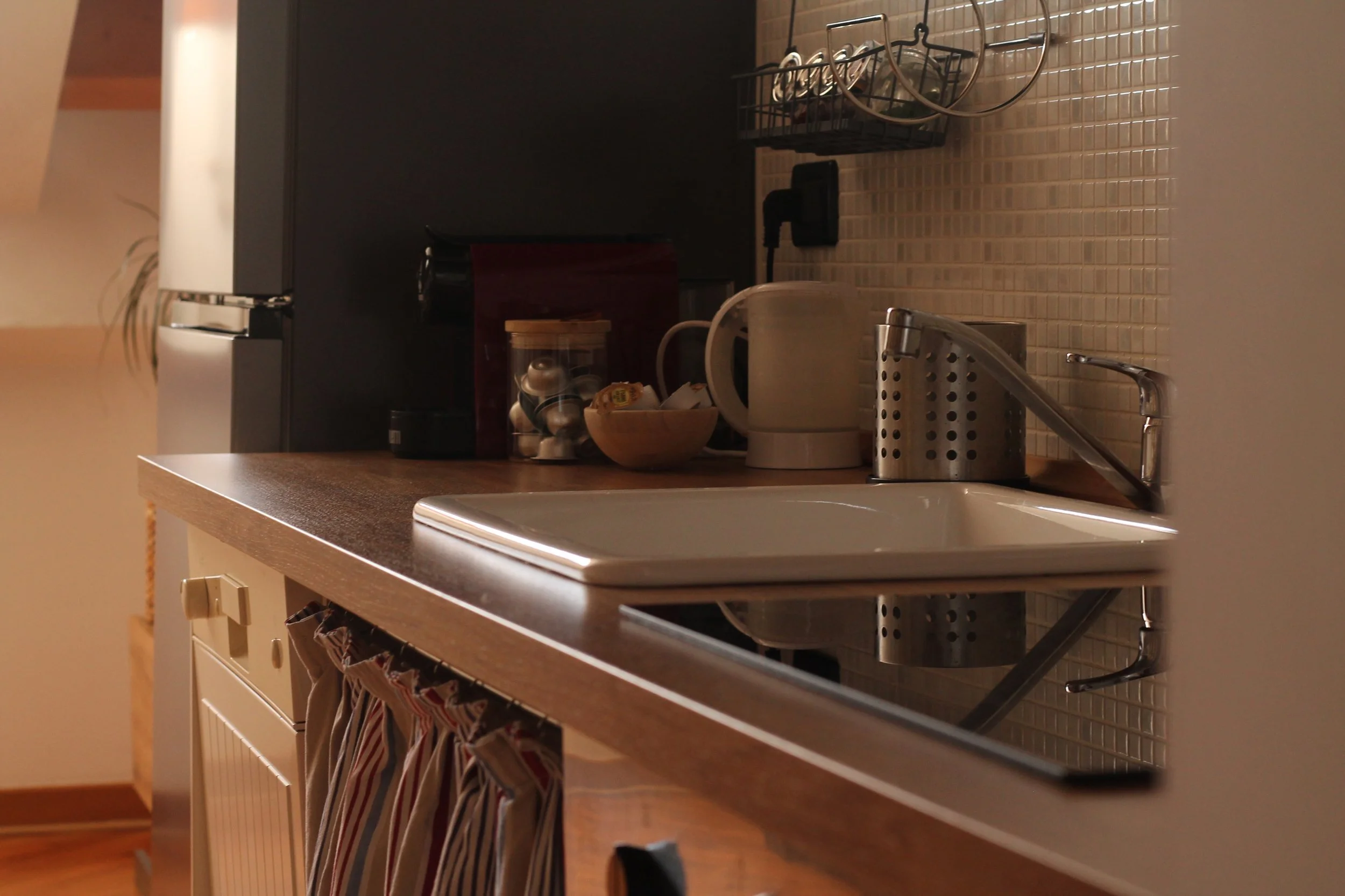 Angolo di cucina con lavabo, estrattore, dispenser, e utensili da cucina sul piano di lavoro, con tendine decorativi sotto il bancone.