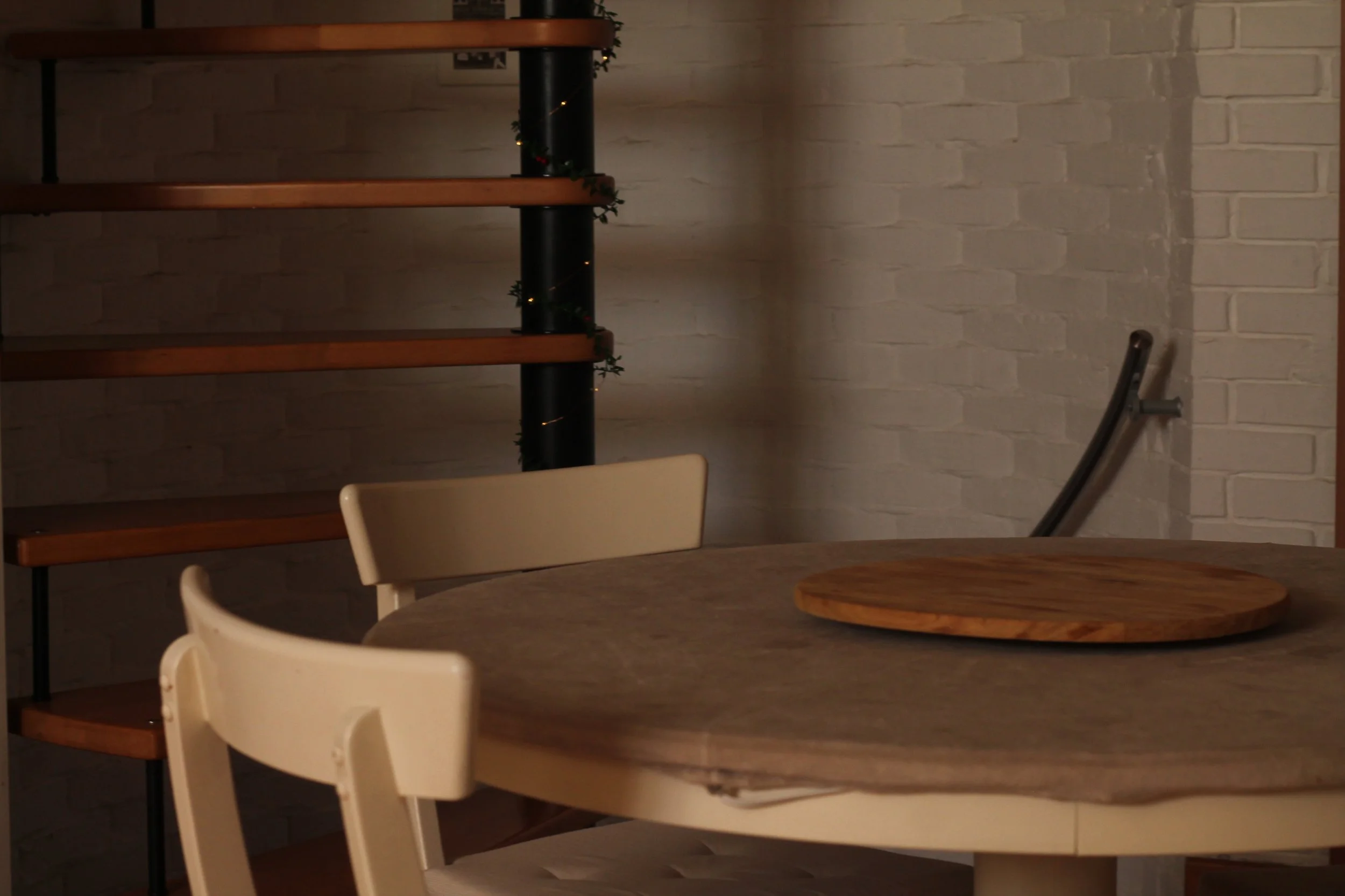 Interno di un ristorante o caffetteria con tavolo rotondo e sedie, parete di mattoni bianchi, scaffale di legno e supporto per Christmas. Fonti di luce soffusa.