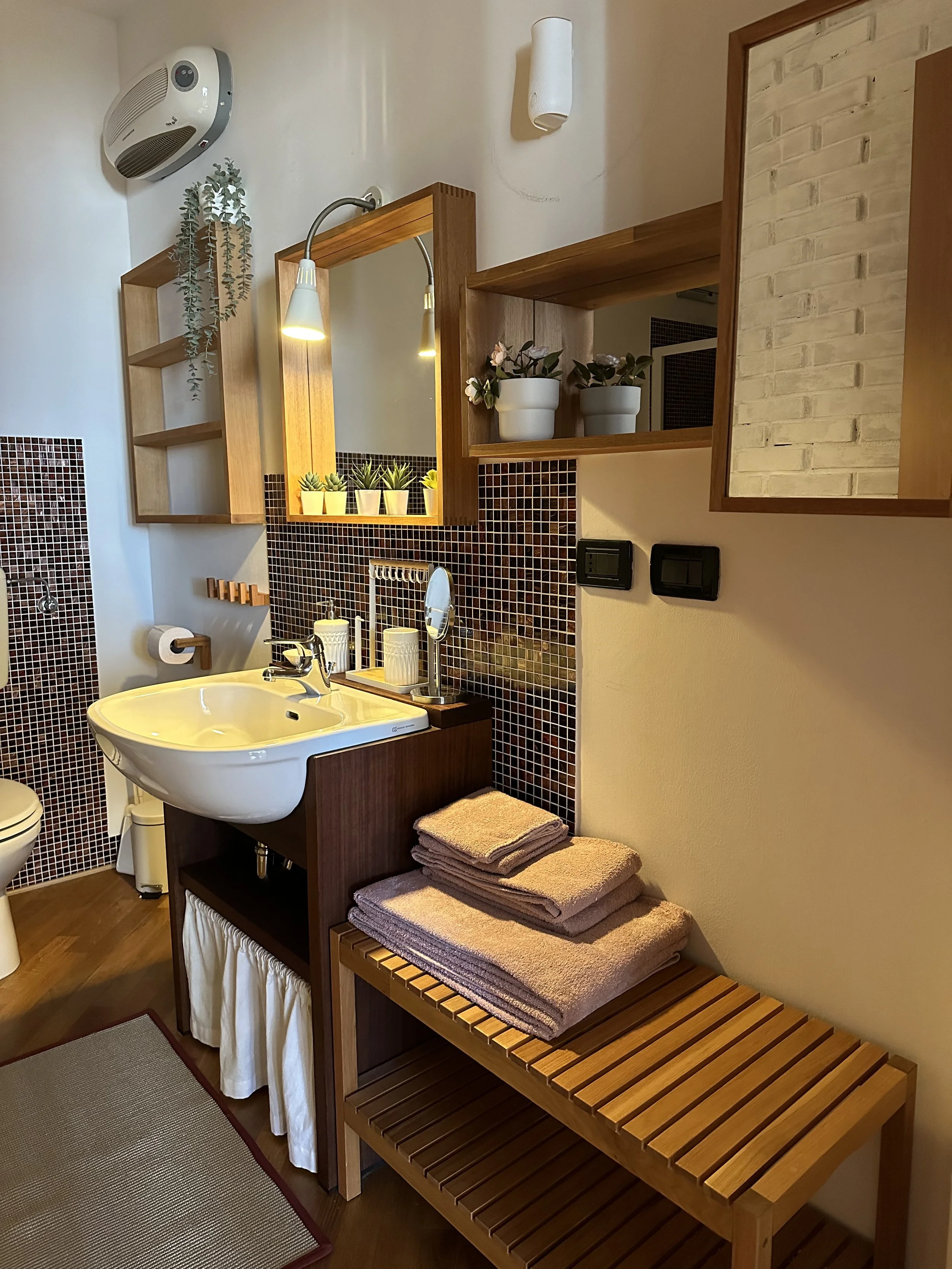 Bagno con lavabo di ceramica bianca su mobile in legno scuro, specchio con cornice di legno, scaffali di legno con piante in vaso, asciugamani impilati su sedia di legno, pareti piastrellate con mosaico in toni marrone e una parete di mattoni bianchi