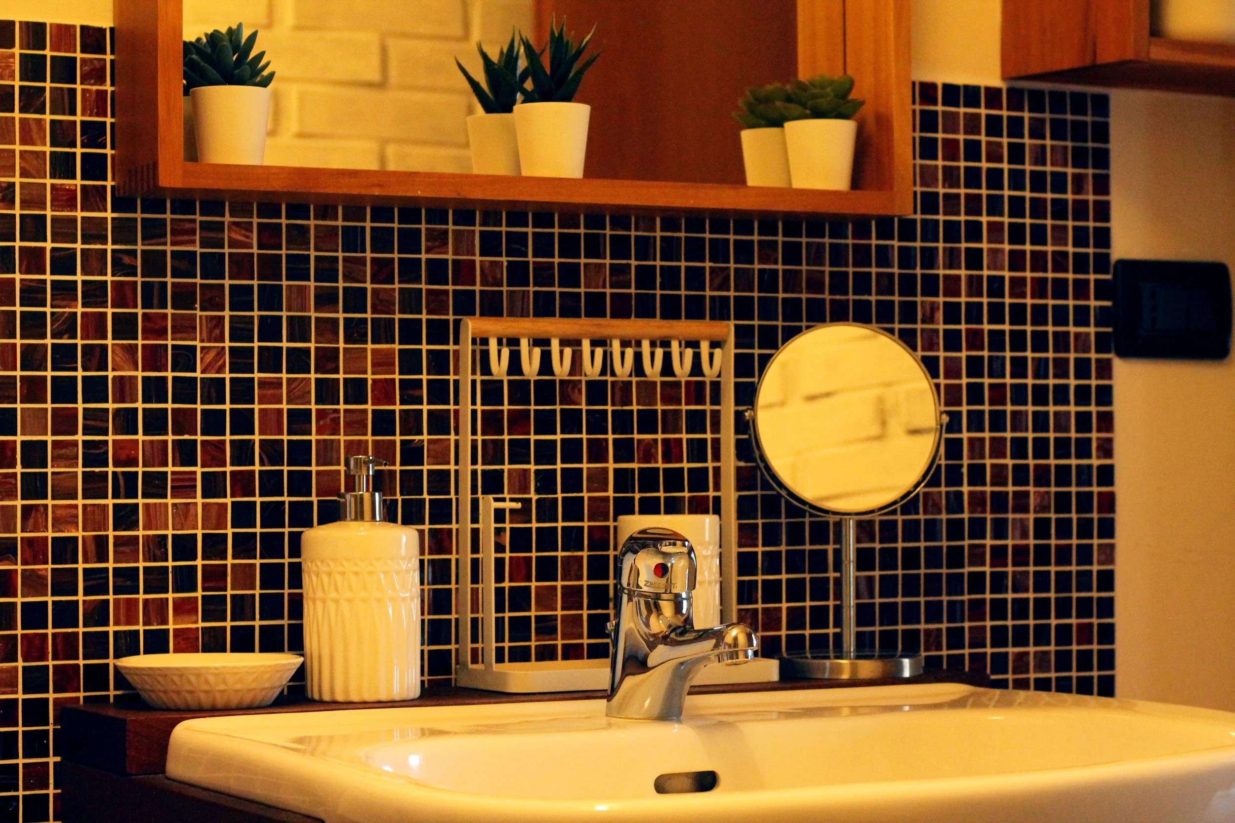 Lavabo con dispenser di sapone, specchio rotondo, pianta e tazzina di ceramica, sullo sfondo piastrelle mosaico colorate