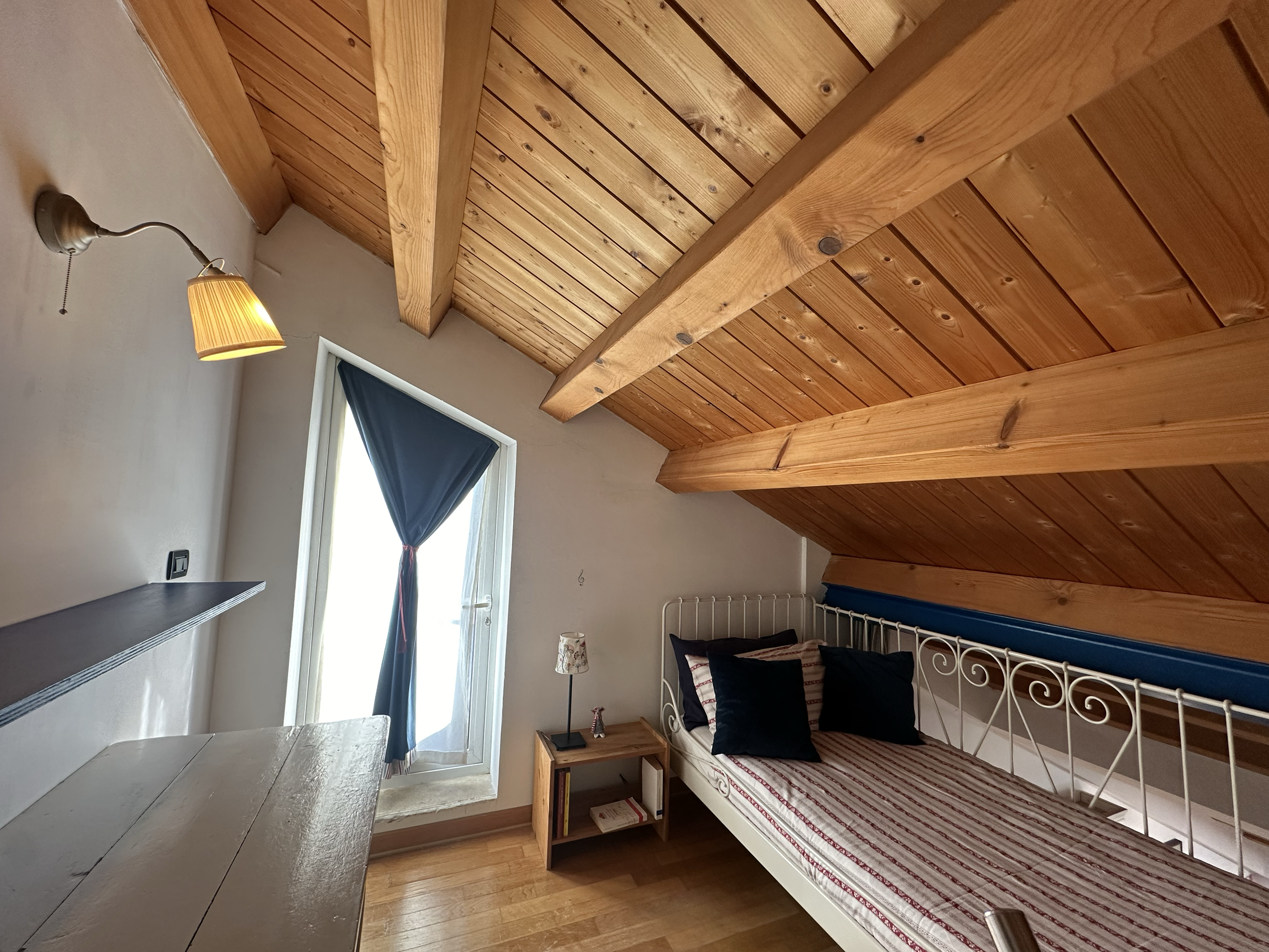 Stanza con letto singolo, pareti bianca, soffitto in legno con travi, finestra con tendina blu, lampada a muro, tavolo in legno, lampada da tavolo, e sedia.