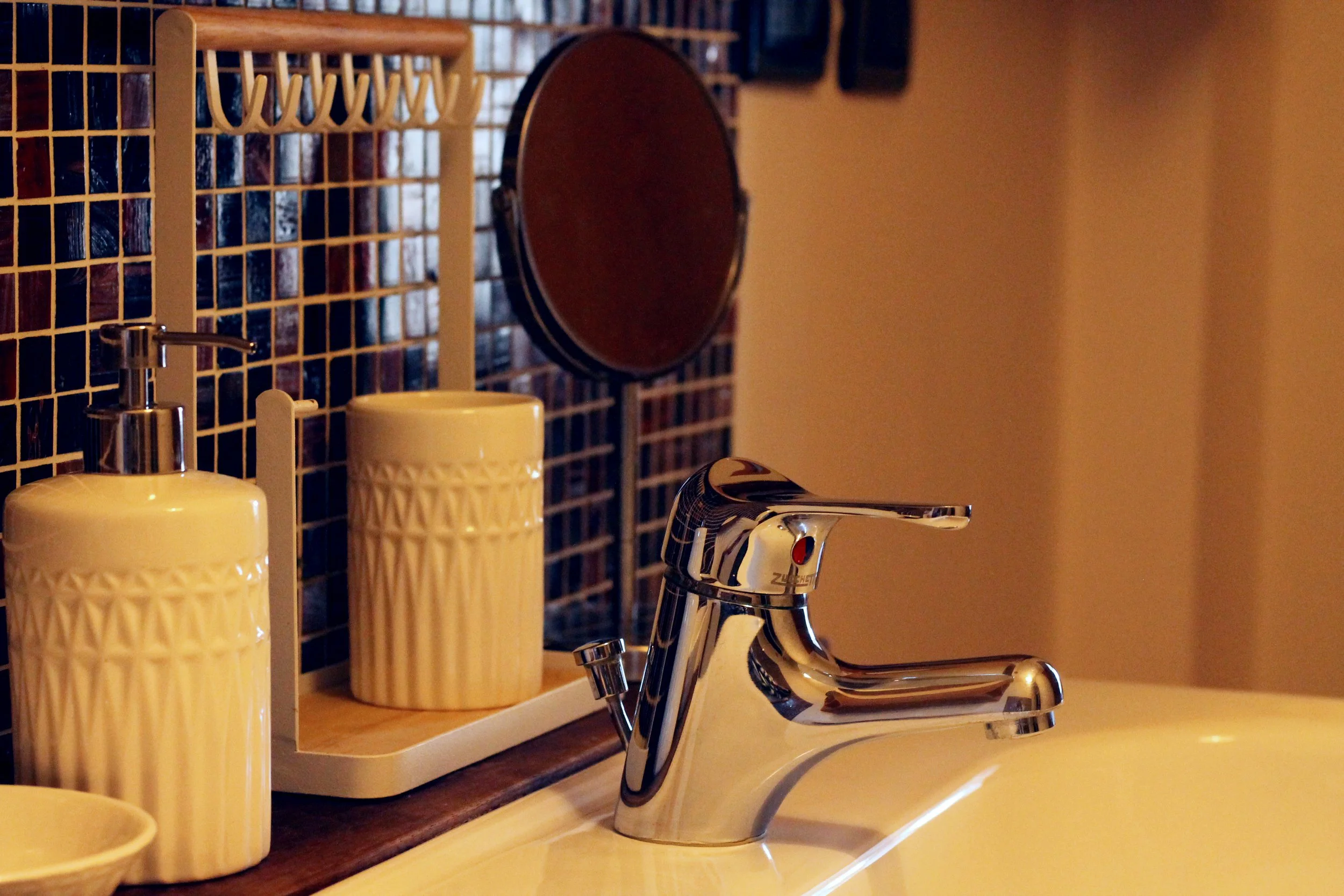 Lavabo con rubinetto cromato, dosatori di sapone e crema in contenitori di ceramica crema, specchio rotondo e mosaico blu sullo sfondo in un bagno.