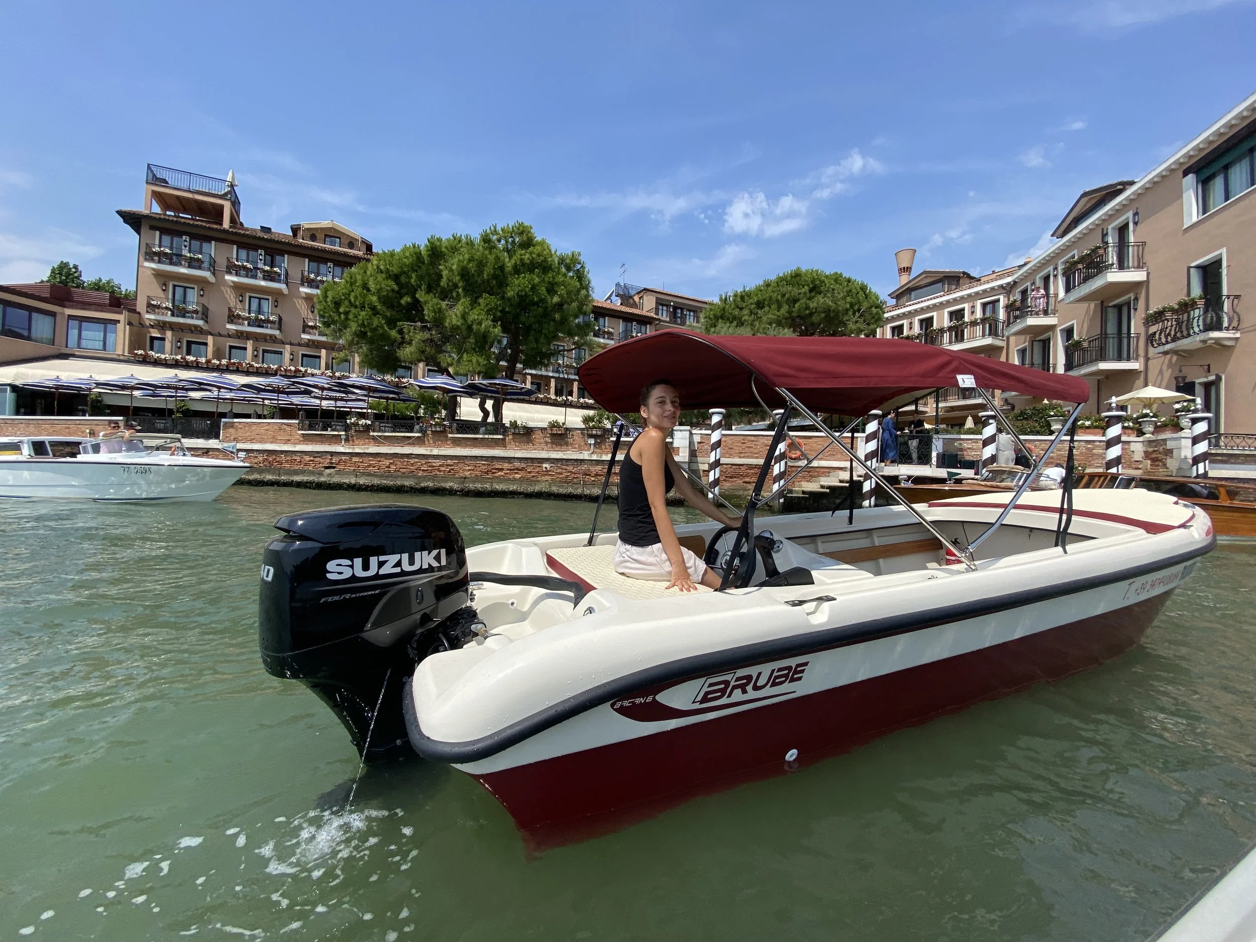 Donna seduta in una barca con motore Suzuki, nella laguna veneziana, con edifici residenziali e alberi sullo sfondo.