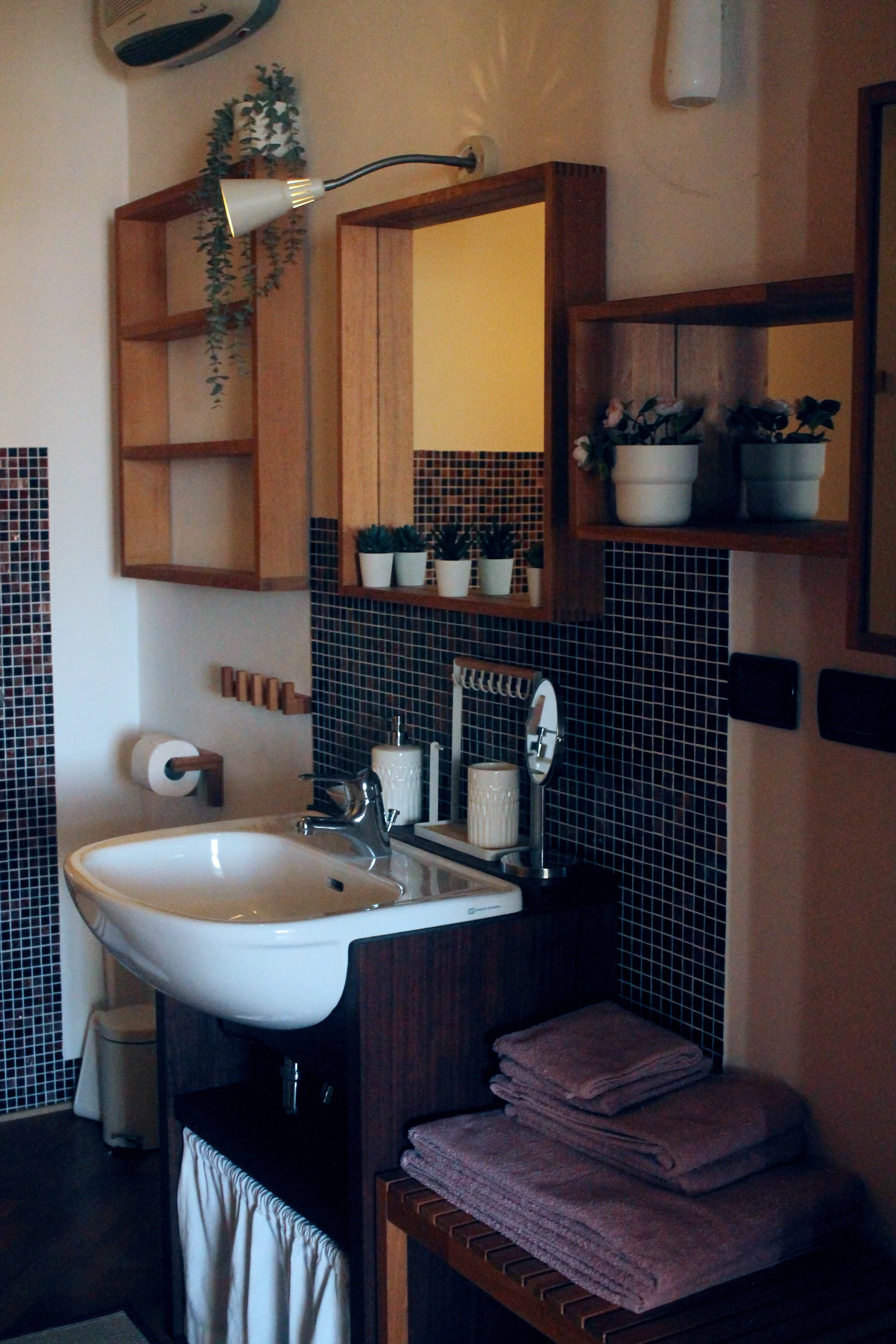 Lavabo con specchio, mensole di legno con piante, asciugamani piegati, carta igienica, utensili da bagno, pareti nere con mosaico.