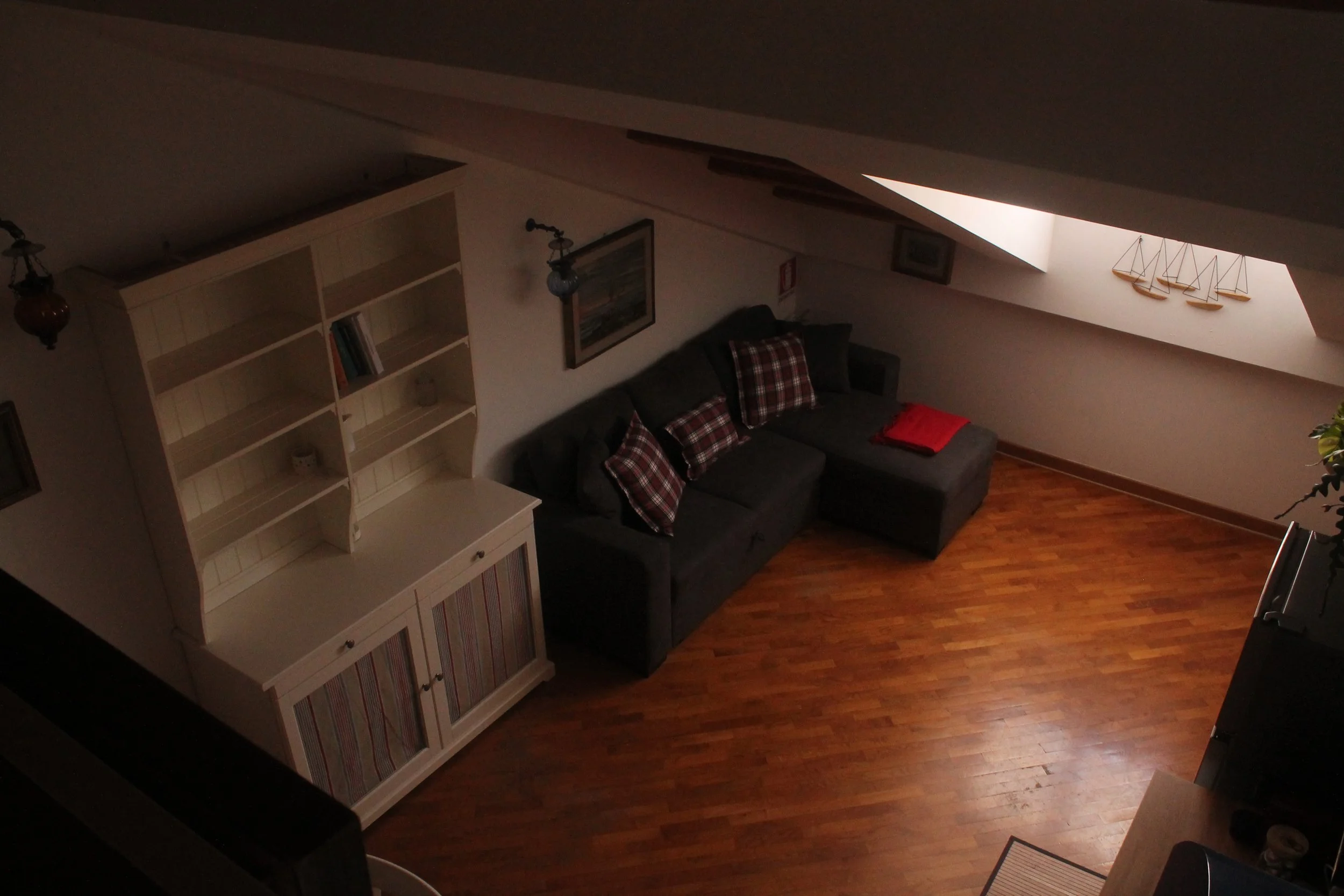 Soggiorno con divano grigio, quattro cuscini con motivo a quadri, parquet in legno, libreria bianca e decorazioni di barche in una finestra velux.