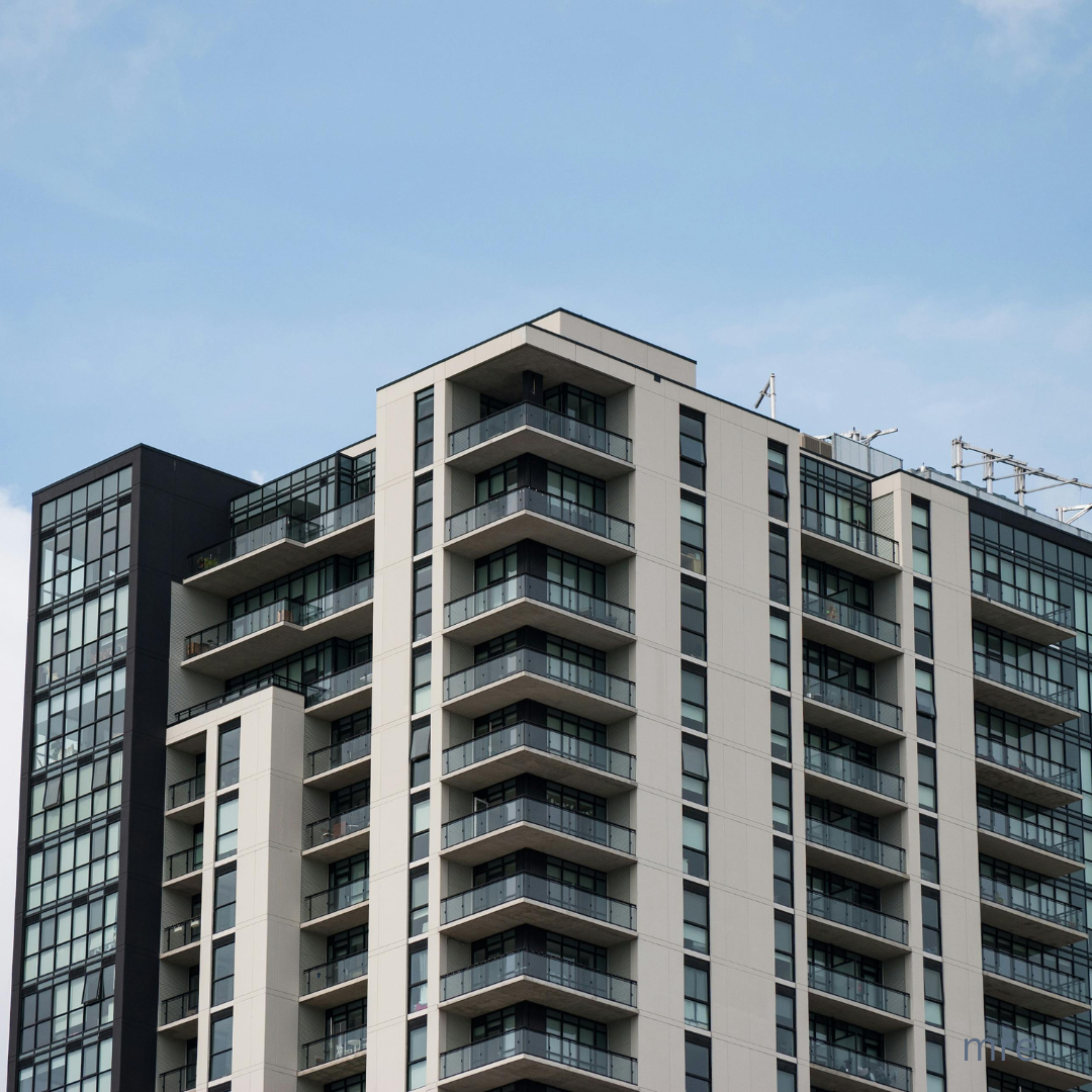 Modern multi-story residential apartment building with multiple balconies, large glass windows, and a clear blue sky background.