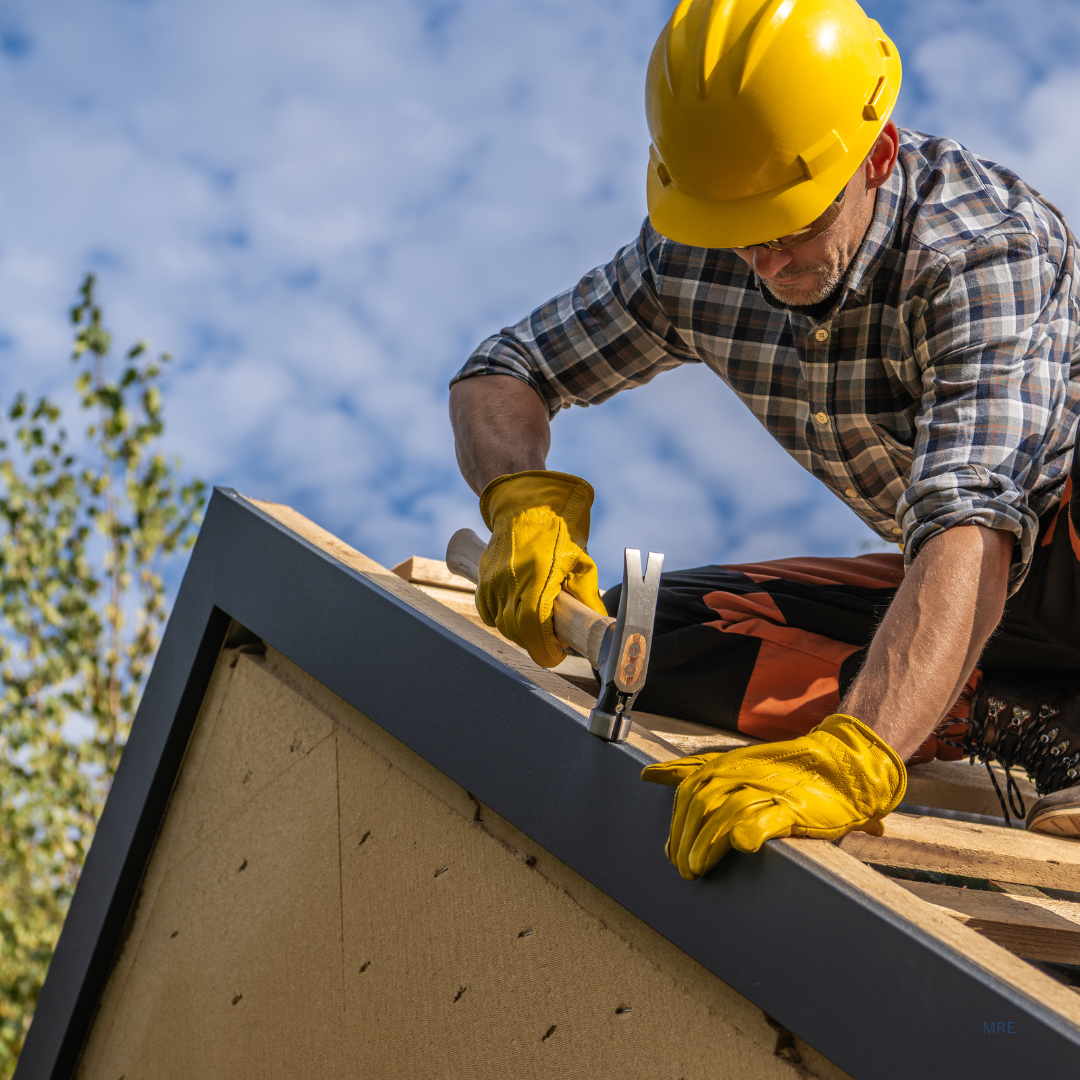 A construction worker wearing a yellow safety helmet and yellow gloves is hammering nails into the edge of a roof on a house under a partly cloudy sky.