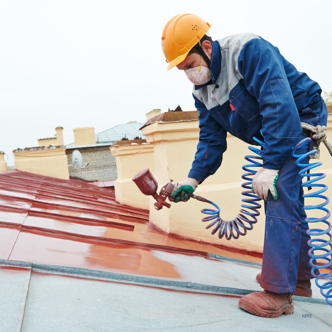 A worker wearing a yellow safety helmet, face mask, and gloves spray-paints a roof red, using a spray gun connected to a coiled hose.
