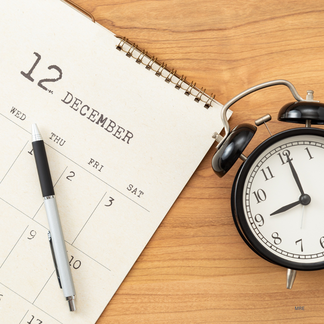 A desk with a spiral calendar open to December 12th, a black and silver pen, and a black alarm clock showing 11:57, on a wooden surface.