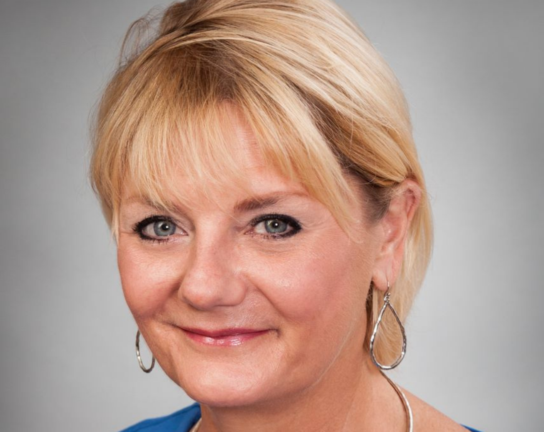 Close-up portrait of a middle-aged woman with short blonde hair, wearing hoop earrings and a blue top, smiling against a gray background.