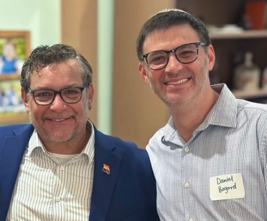 Two men with glasses smiling at the camera in an indoor setting. One is wearing a blue blazer with a small American flag pin, and the other is wearing a striped shirt with a handwritten name tag that says 'Daniel Bagard'.