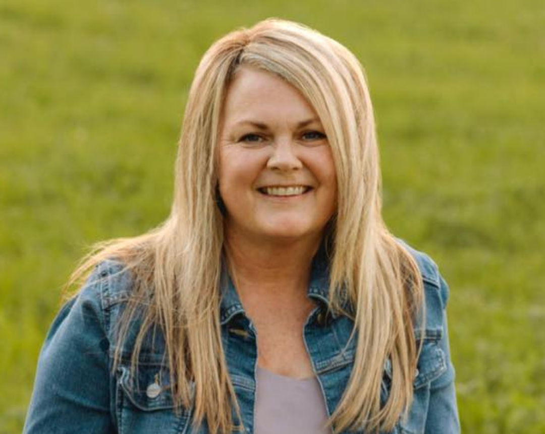 Smiling woman with long blonde hair wearing a denim jacket outdoors with green grass in the background.