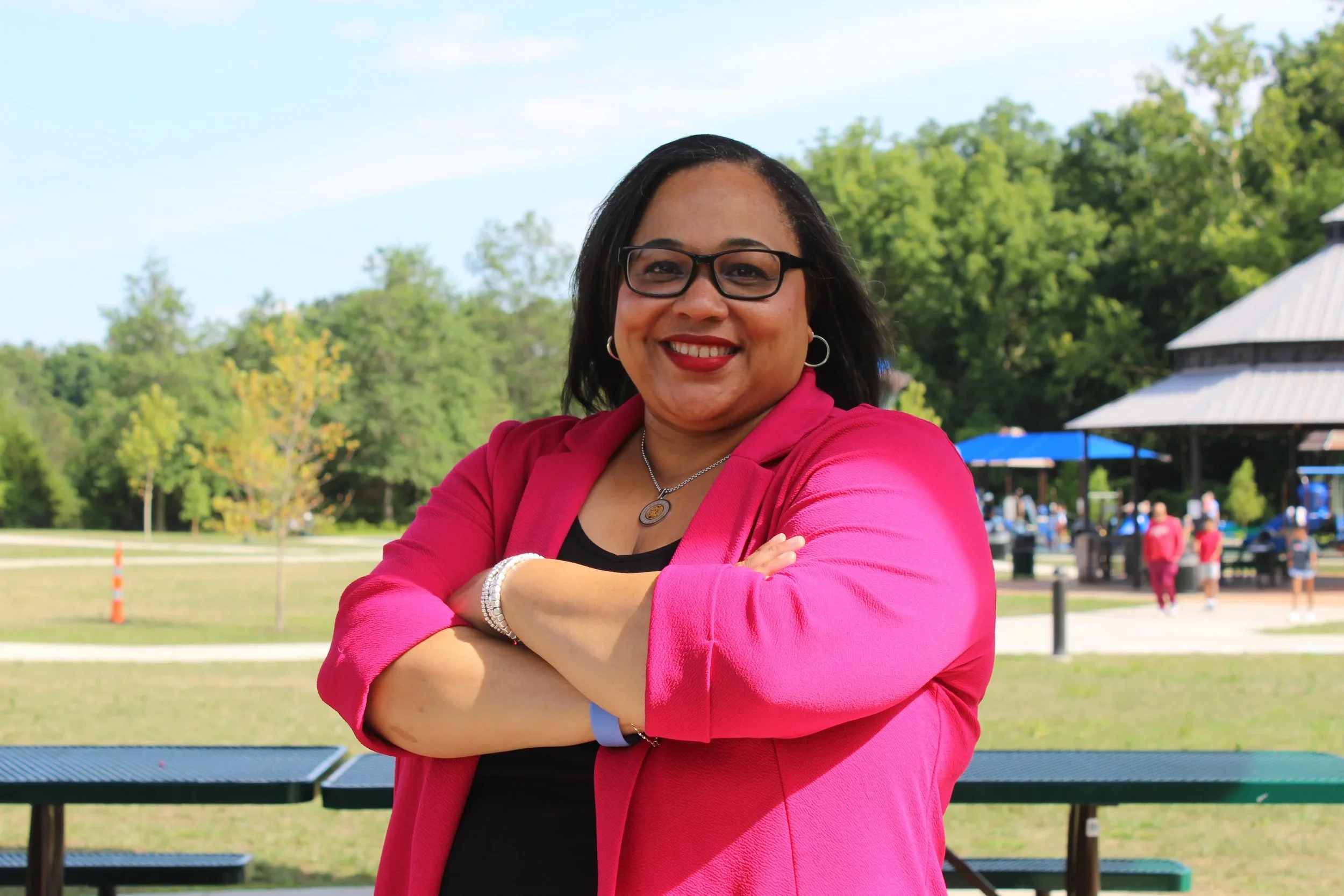 A woman with glasses and hoop earrings smiling with arms crossed, wearing a pink blazer and black top, outdoors in a park with trees, a pavilion, and people in the background.