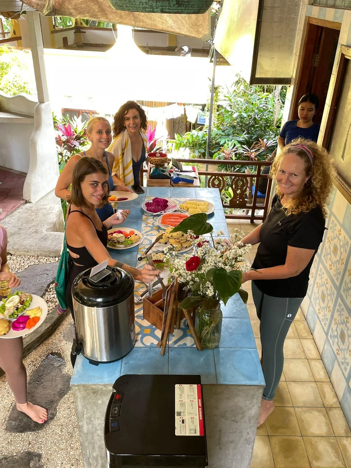 A group of five women at a table set with plates of food, including sandwiches and fruit, in a bright, open-air setting with plants and decorative tiles, with a woman standing and four women seated, smiling at the camera.