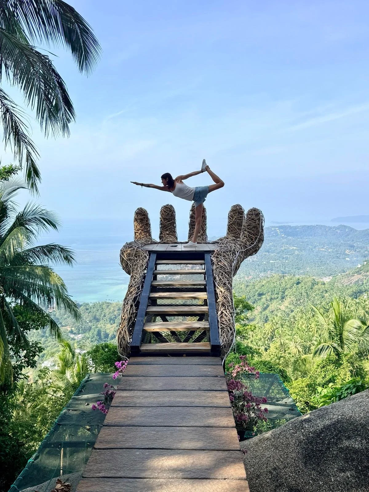 A woman practicing yoga in a handstand pose on a giant wicker hand-shaped sculpture on a mountain overlooking a lush green landscape and ocean.