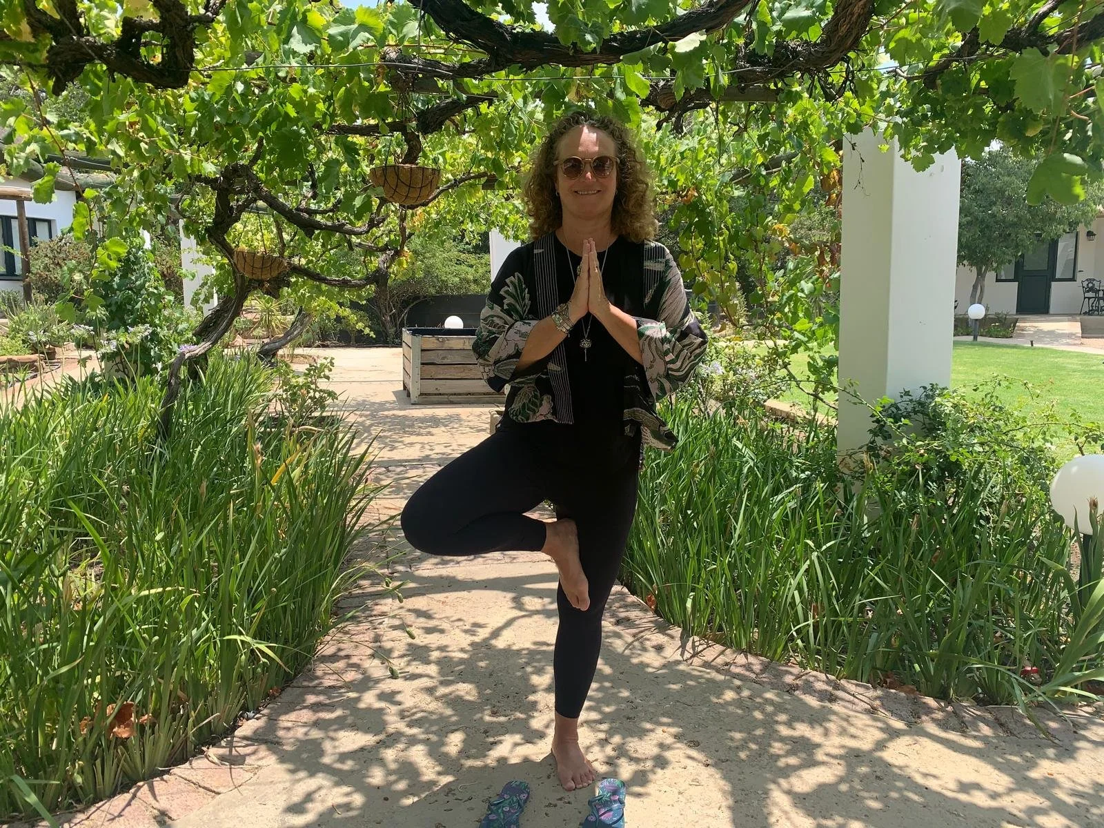 Woman practicing yoga in a garden, standing on one leg with hands in a prayer position, surrounded by greenery and shaded by a vine-covered pergola.