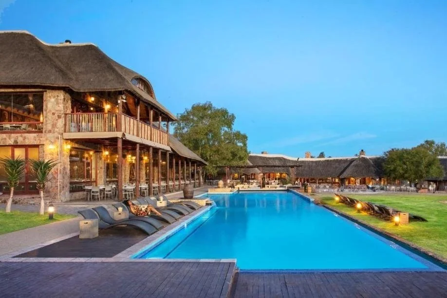 Luxury resort with a large swimming pool, poolside lounge chairs, a two-story thatched-roof building, outdoor dining area, and landscaped grass and trees at dusk.