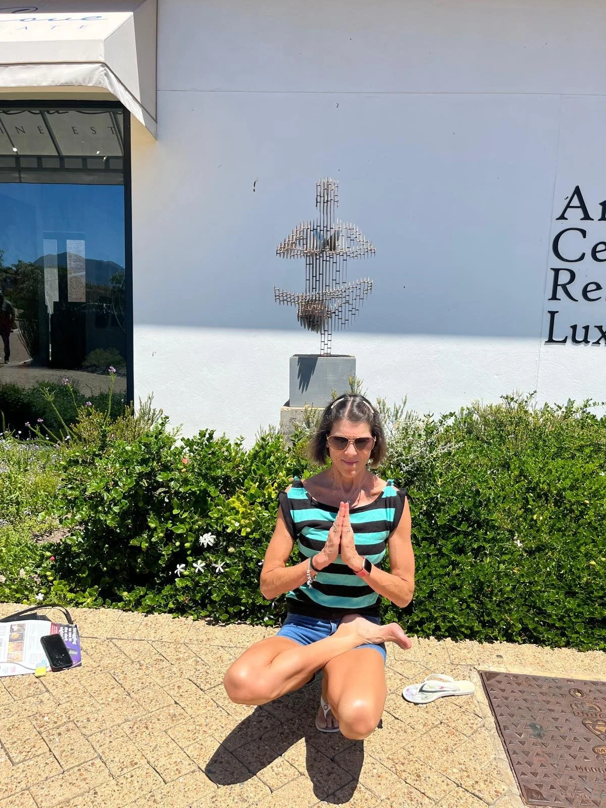 A woman with short hair, sunglasses, and a striped shirt is doing a yoga pose outdoors in front of bushes, with her hands in prayer at her chest. She is kneeling on one knee and has a calm expression. There are personal items on the ground, and a sculpture of a human figure made of metal bars on a pedestal behind her. The scene is beside a white building with black lettering and some greenery.