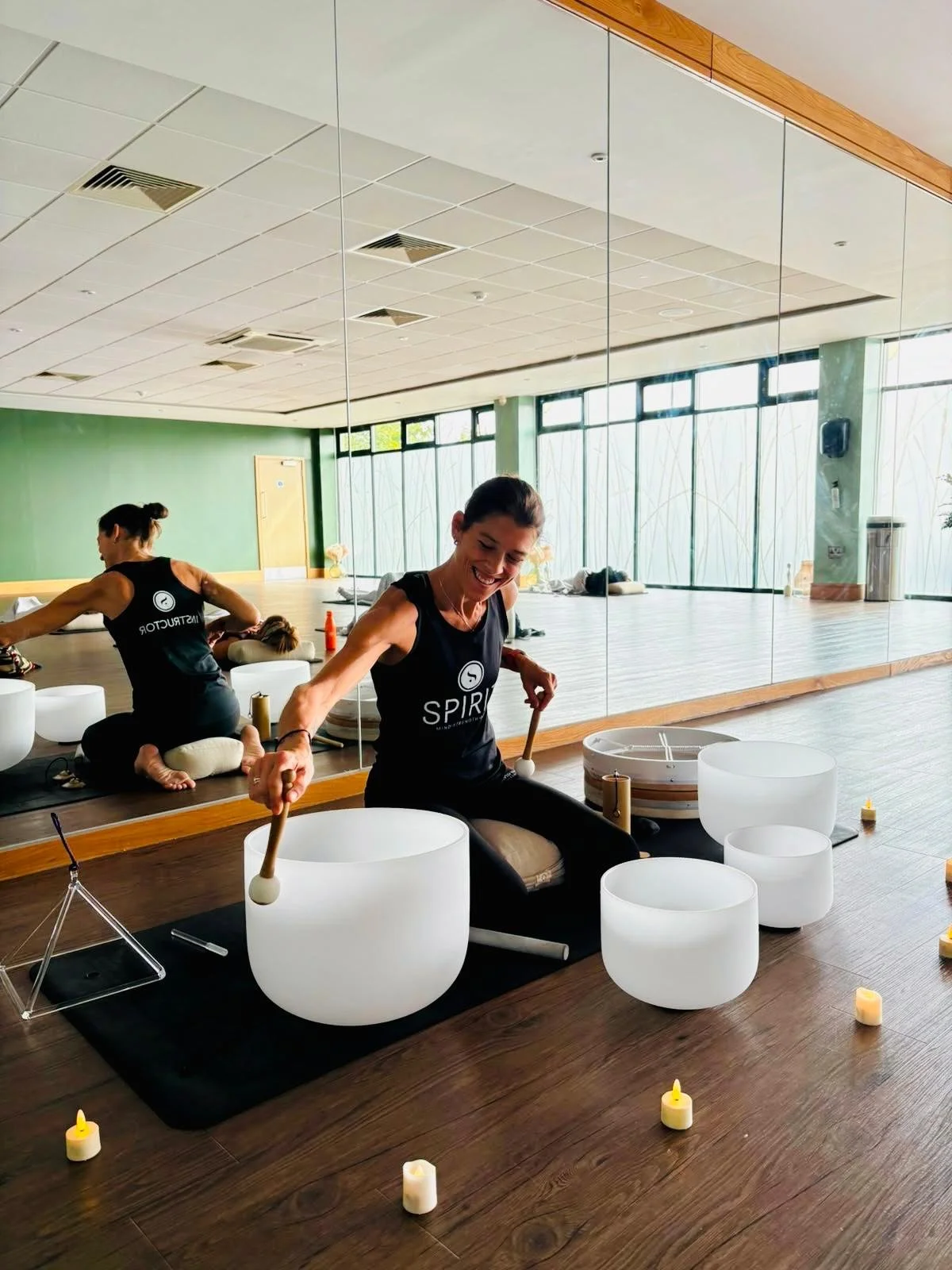 A woman sitting cross-legged, playing crystal bowls in a yoga or meditation studio with candles arranged around her.