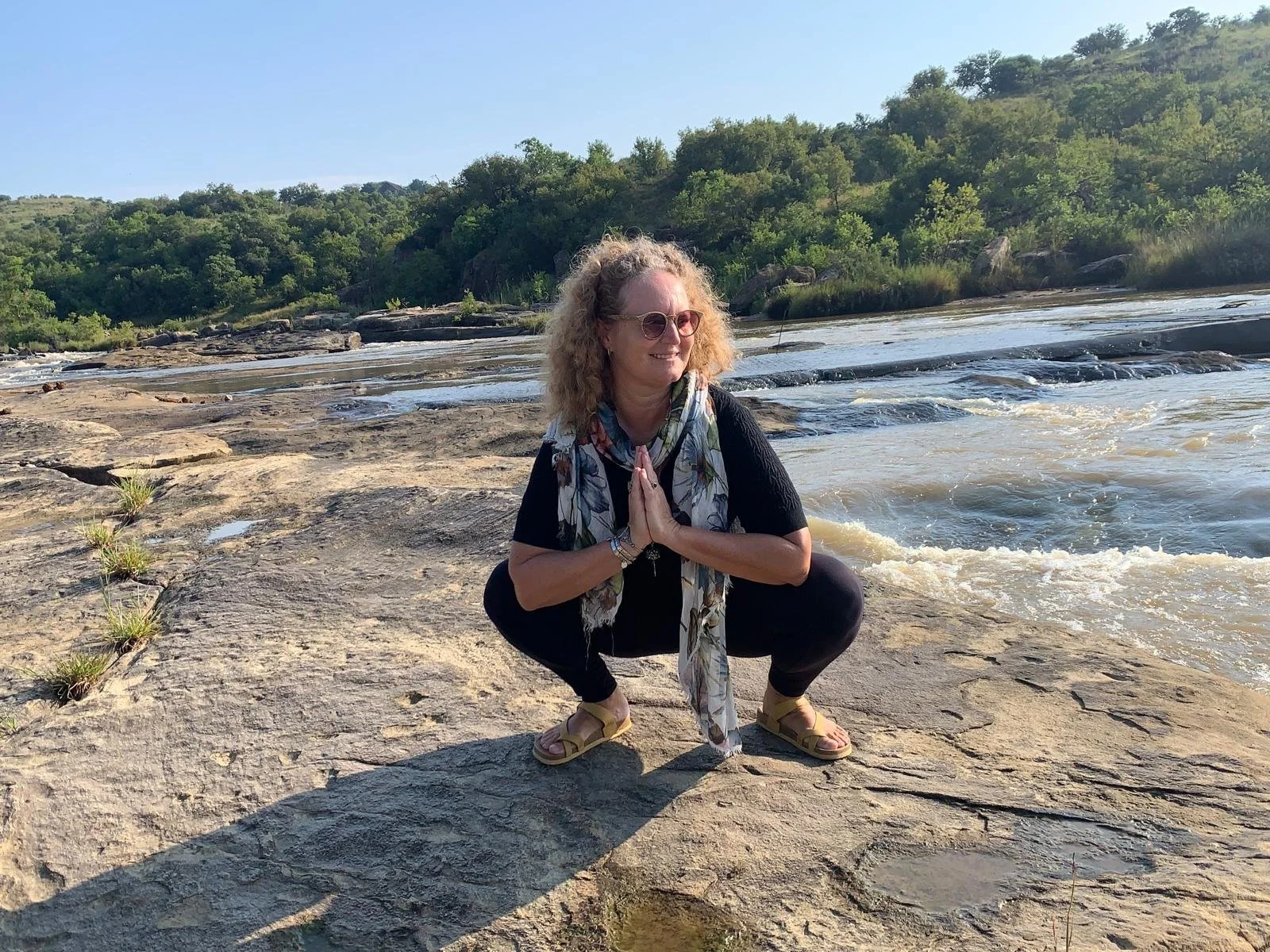 Woman with curly blonde hair wearing sunglasses, black shirt, and a colorful scarf practicing yoga or meditation by a river with green hills and trees in the background.