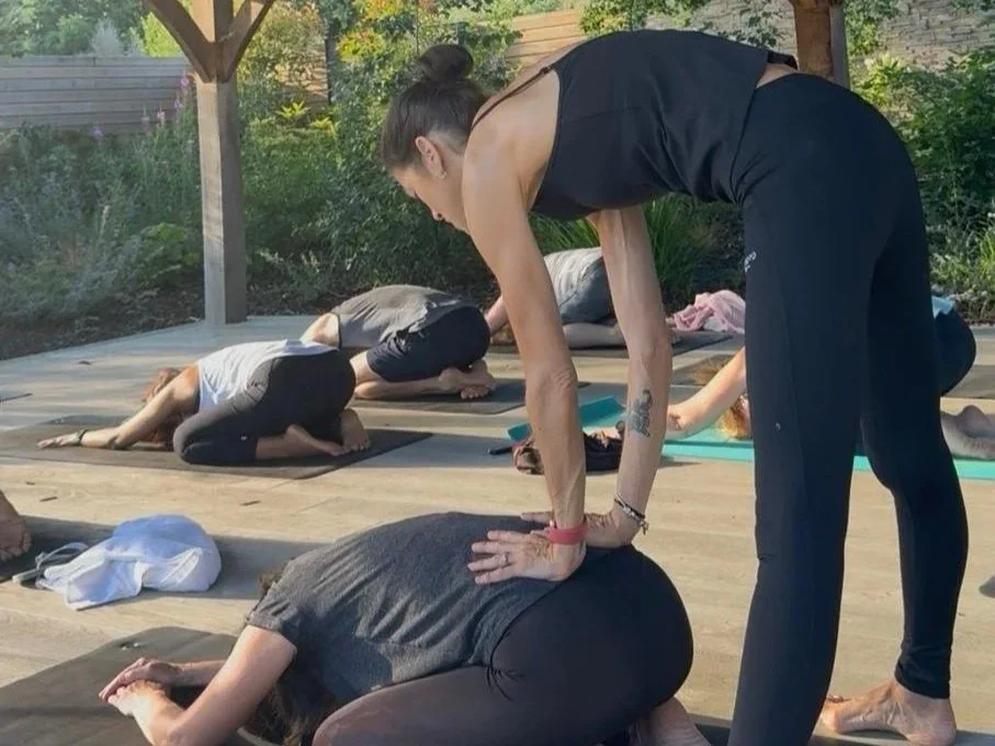 A woman leading a yoga class outdoors, demonstrating a forward bend pose to a participant.