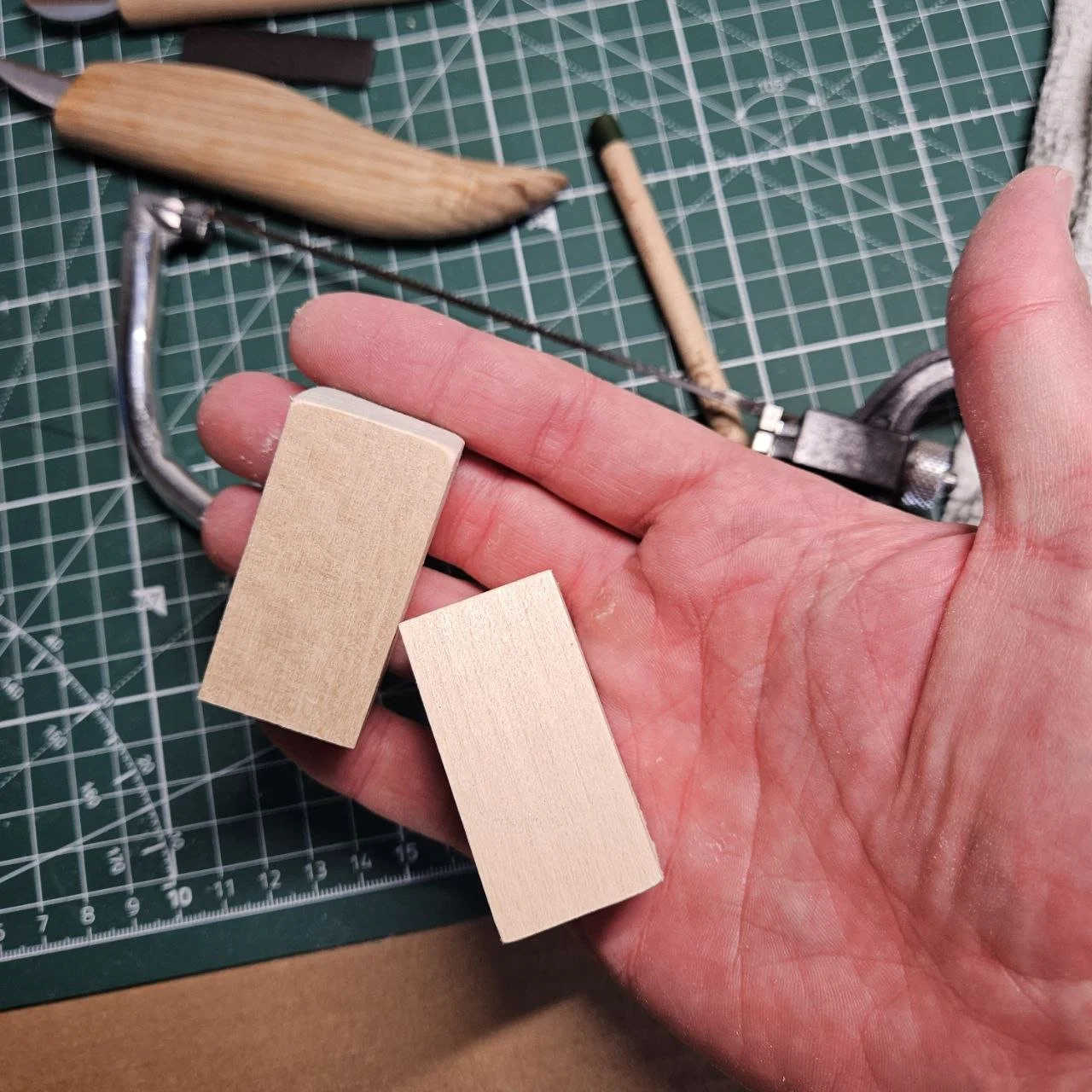 Due pezzi di legno chiaro e uno strumento per tagliare il legno afferrati da una mano, con un tavolo da lavoro con strumenti e materiali.